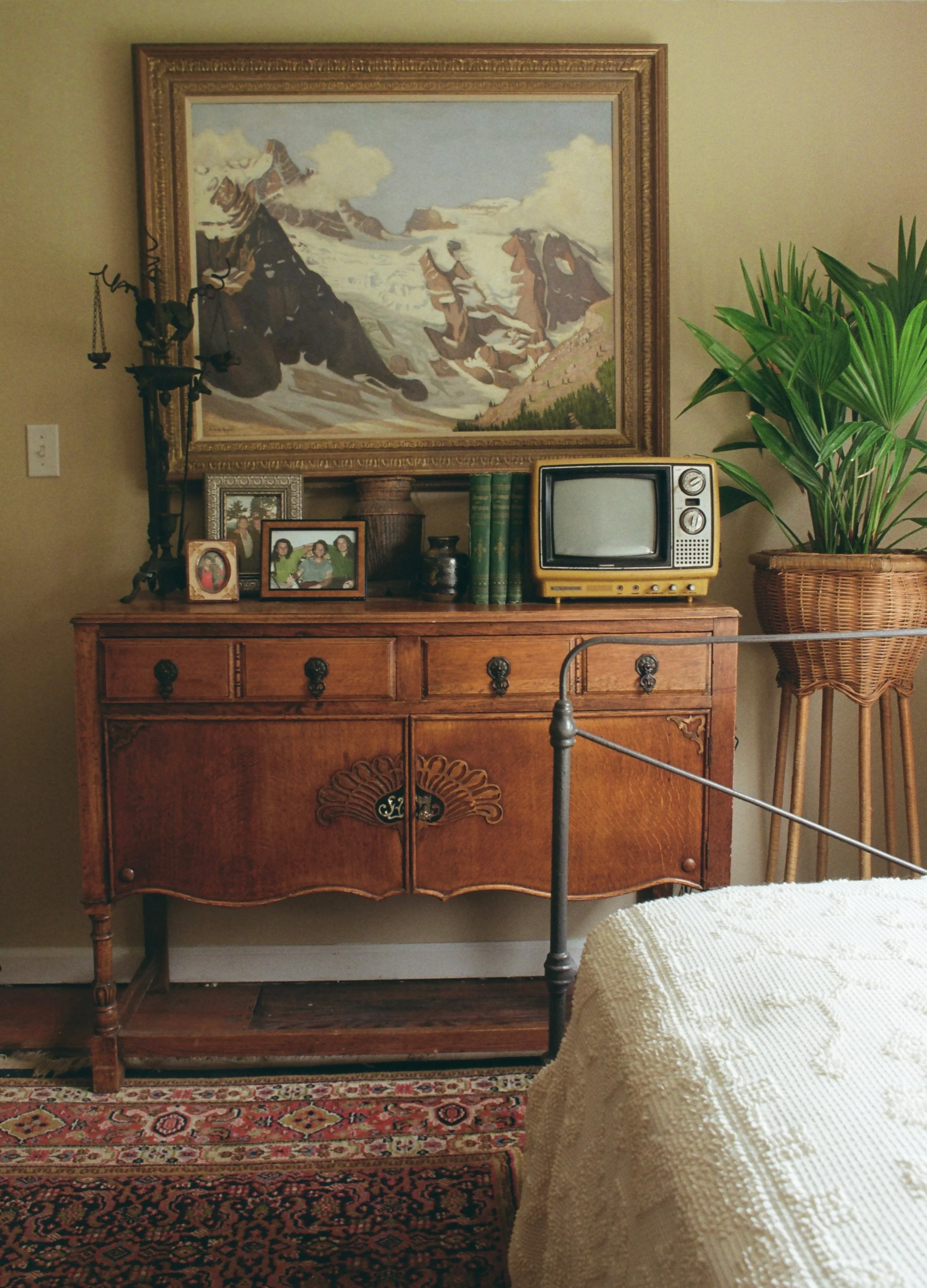 A wooden antique dresser with ornate handles beneath a large framed painting of a snowy mountain landscape. The dresser holds family photos, a small framed picture, a set of three green books, a vintage TV, and decorative vases. To the right, there is a large potted plant with long green leaves, and part of a wicker chair with a textured cream-colored cushion is visible.