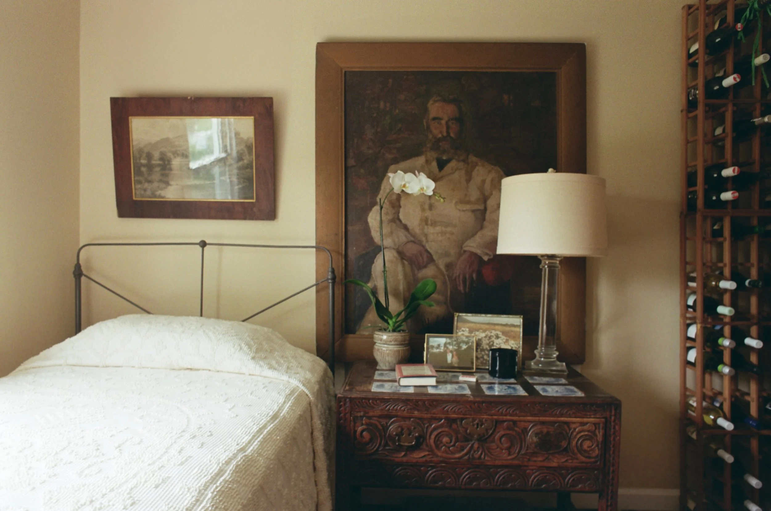 A bedroom corner with a bed covered in a white textured blanket, a wooden bedside table with framed photos, a white orchid plant, a table lamp with a white shade, a large framed portrait of a man with a beard, a smaller framed picture, and a wooden wine rack filled with bottles.