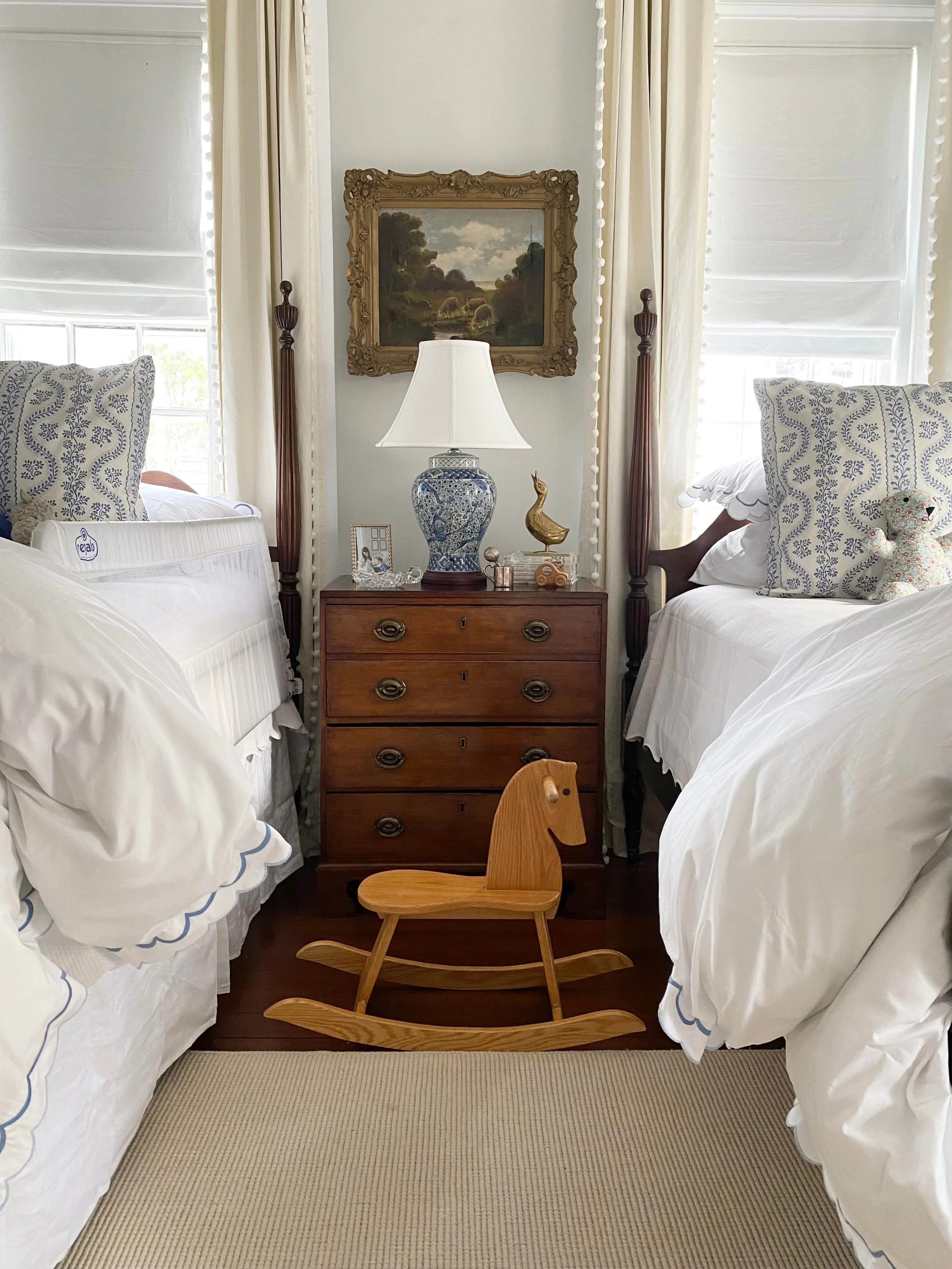 A bedroom with two twin beds with white bedding and blue trim, a wooden rocking horse in between, and a wooden chest of drawers with a blue and white lamp, decorative items, and framed photos. There are windows with white blinds and curtains, a framed landscape painting on the wall, and beige carpeted floor.