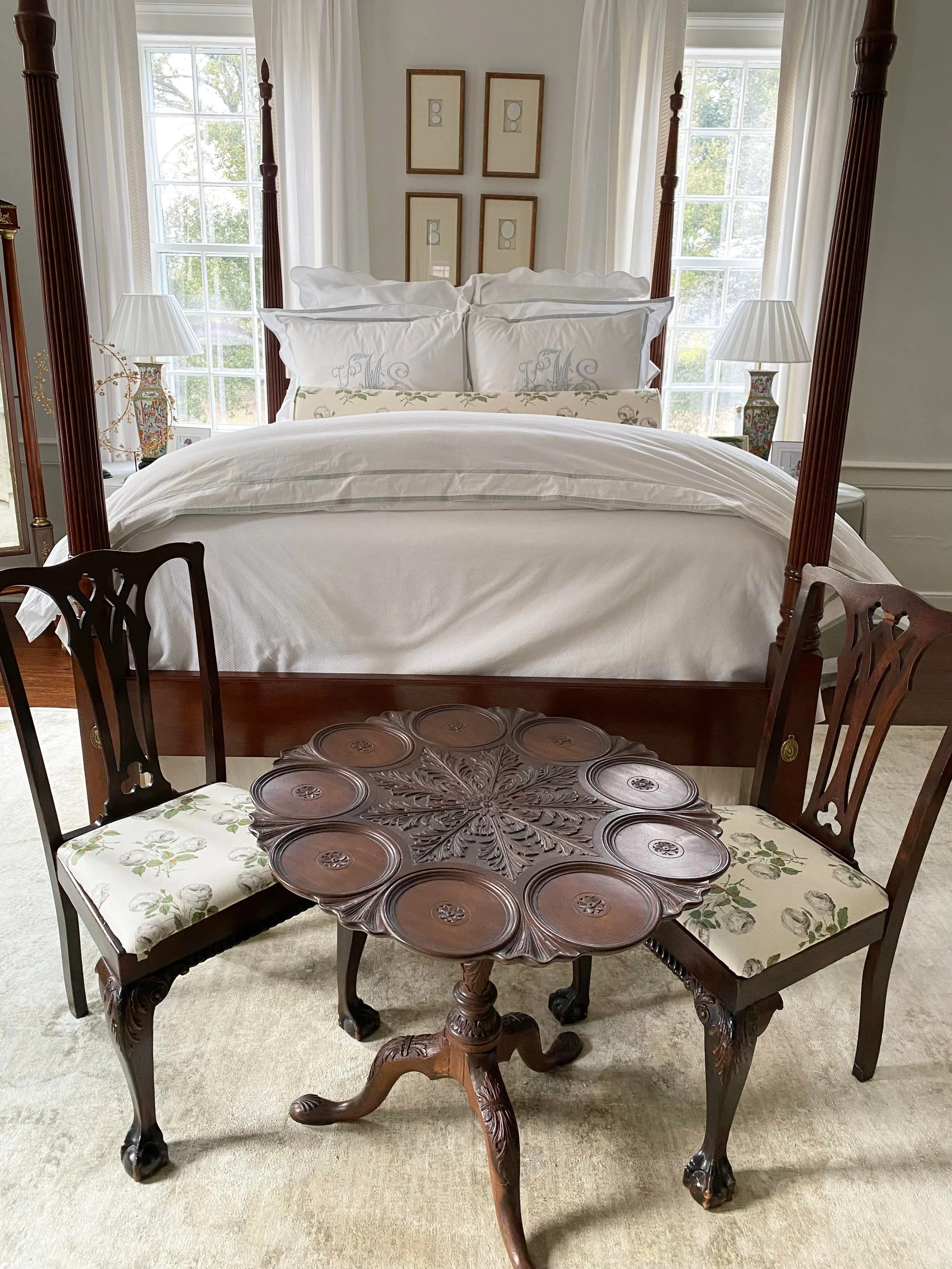 A bedroom with a four-poster bed with white bedding, two nightstands with lamps, and window blinds. A round wooden table with carved details and four chairs is in front of the bed.