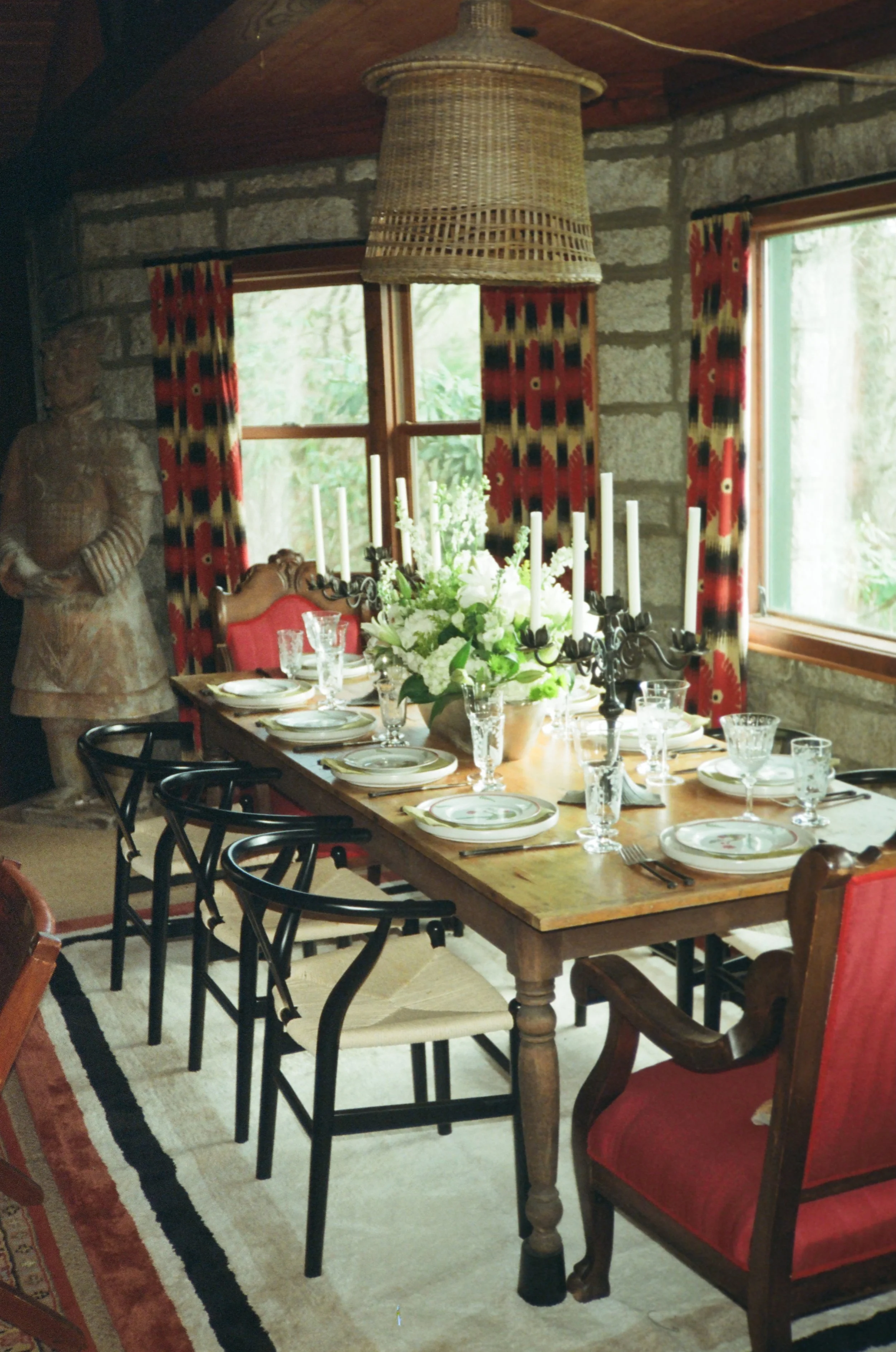 A dining room with a wooden table set with plates, glasses, and a floral centerpiece, surrounded by various chairs, featuring stone walls, red and black patterned curtains, and a large wicker lamp hanging from a wooden ceiling.