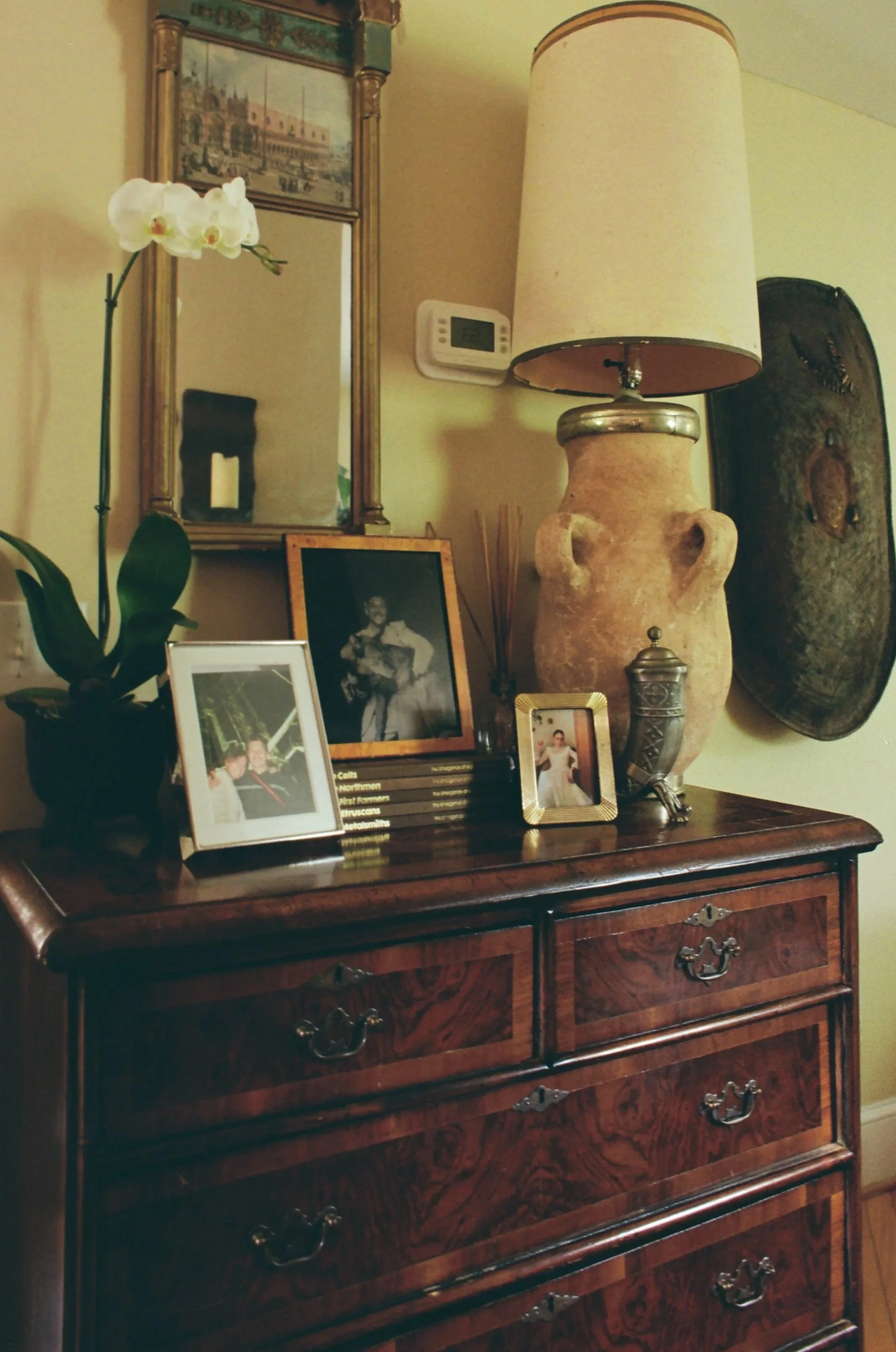 Decorative wooden dresser with brass handles featuring framed photographs and a large ceramic urn with a lamp shade on top, situated against a yellow wall with a mirror, a painting, a thermostat, and a large decorative tray in the background.