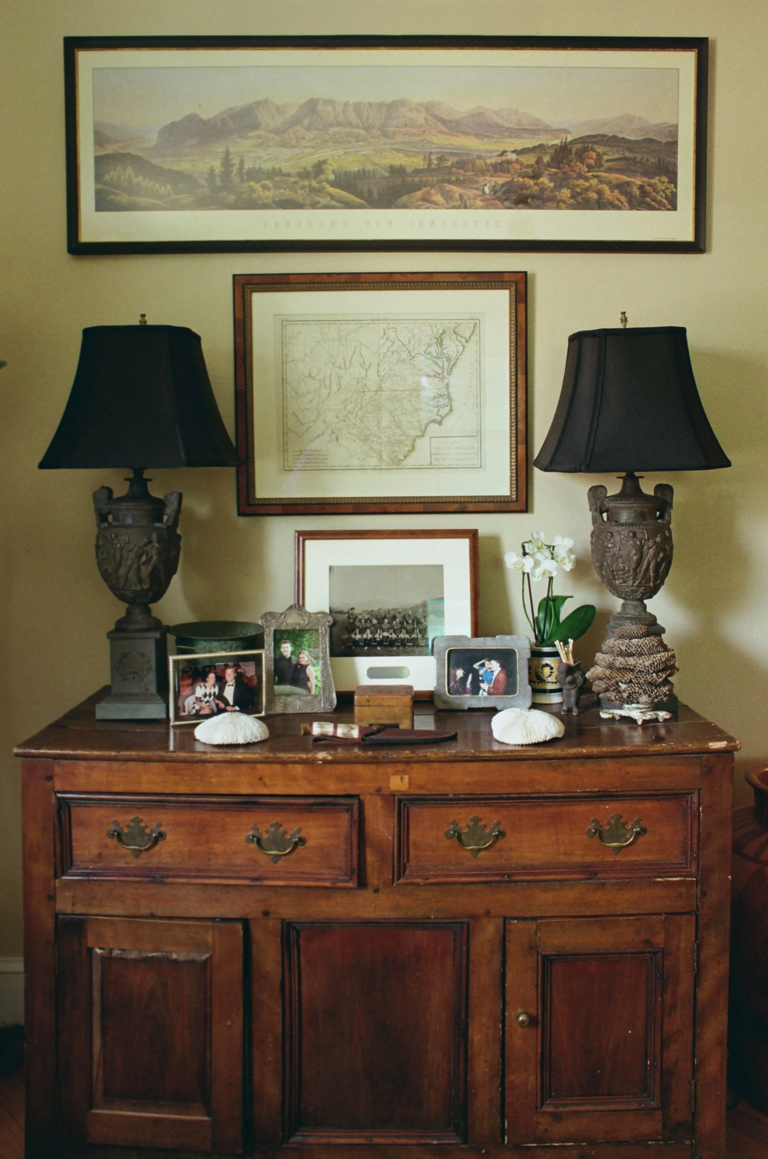 A wooden dresser with two dark lamps with black lampshades on top. The dresser has three drawers with metal handles and two cabinet doors at the bottom. Decorated with framed photos, a small burlap-wrapped object, a plant, and small decorative items. Three wall art pieces hang above: a landscape painting of mountains and green fields, a framed map of Arkansas, and a blank frame below them.