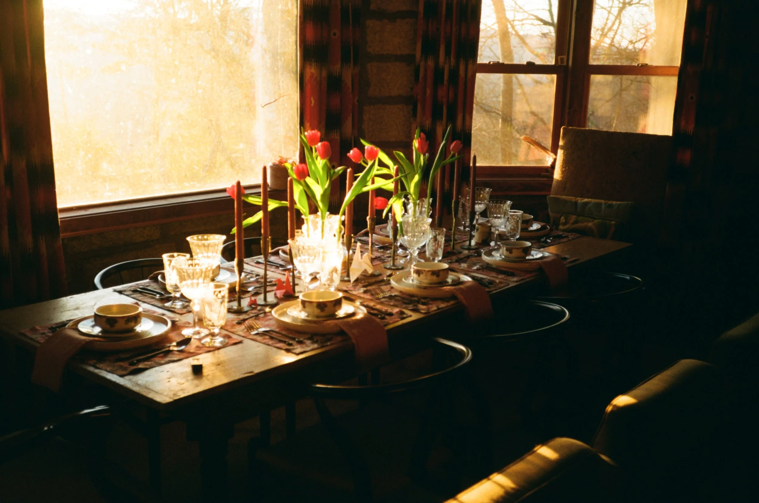 A dining room table set with teacups, glasses, plates, and silverware. A clear glass vase with pink tulips and tall candles is in the center. The room has large windows with natural light and curtains, and there is a chair visible on the right side.
