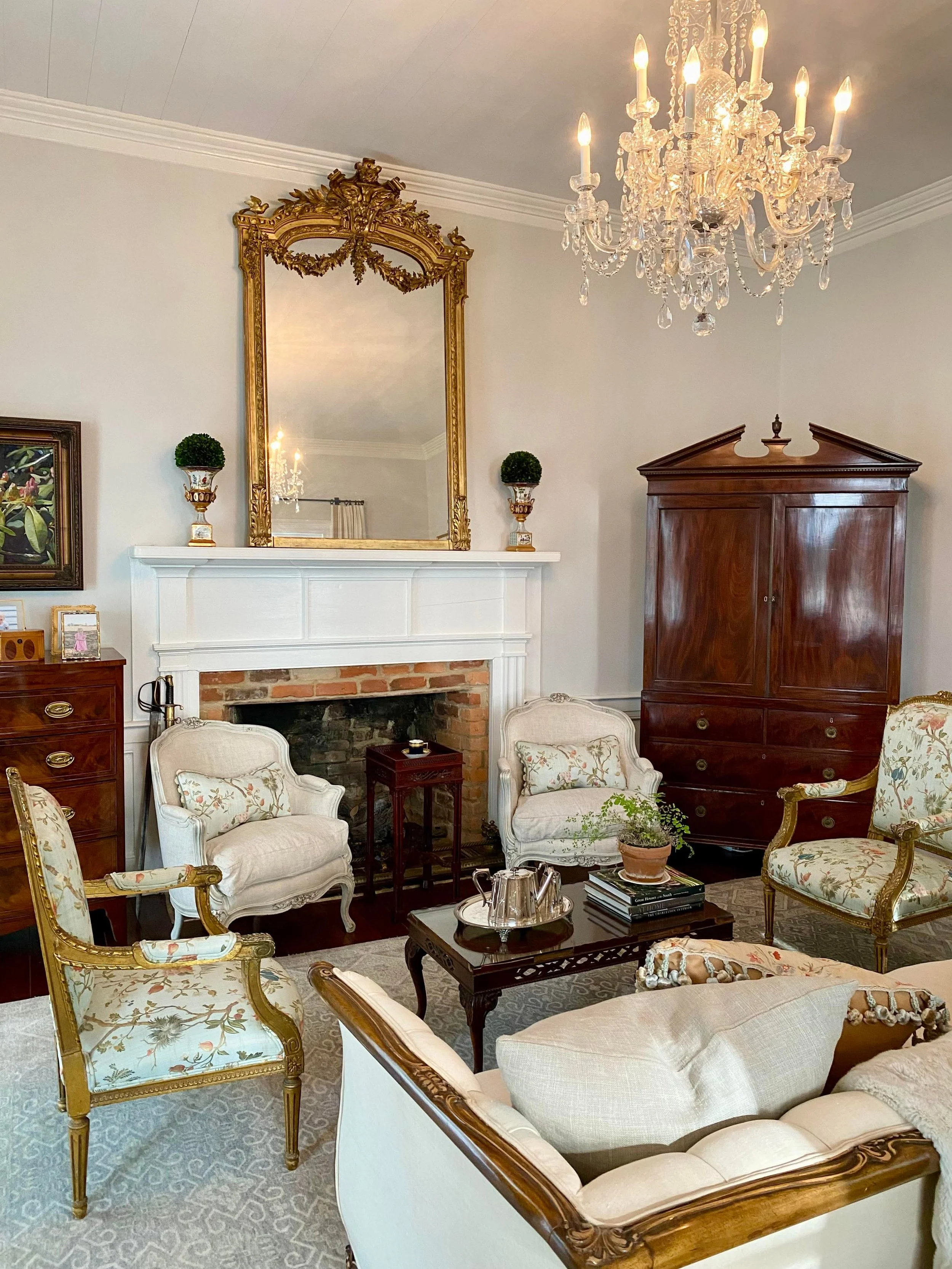 A living room with elegant antique furniture, a white fireplace, a large ornate mirror with a gold frame above the fireplace, a chandelier hanging from the ceiling, potted plants, and picture frames on the mantel.