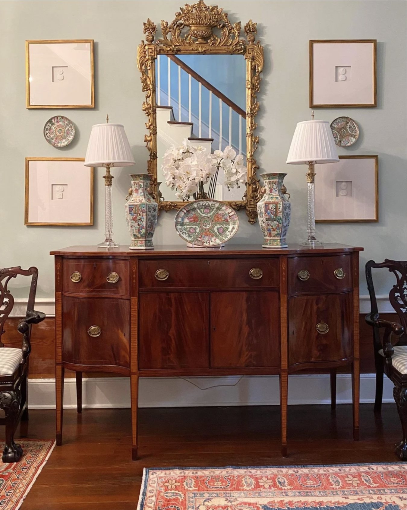 A wooden sideboard with a curved front and four drawers, topped with four decorative vases and a floral arrangement, stands against a pale blue wall. Above it hangs an ornate gold-framed mirror, flanked by two empty square gold picture frames and two small decorative plates. There are two table lamps with white shades on either side of the mirror. A staircase with white railings and wooden steps is reflected in the mirror. A patterned rug with red, cream, and blue colors is partially visible on the floor, and parts of two wooden chairs with patterned cushions are seen on either side of the sideboard.