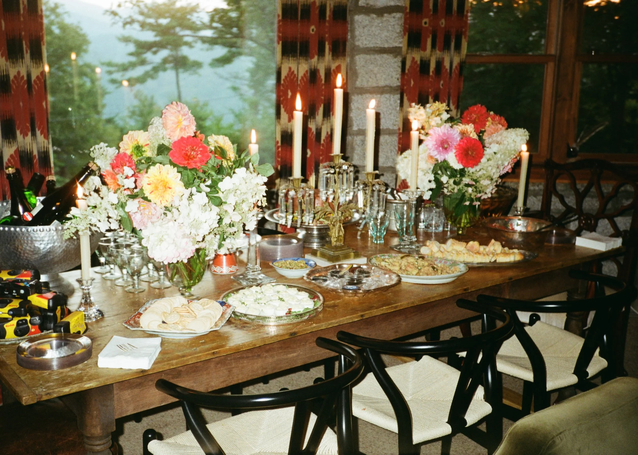 A wooden dining table decorated with multiple flower bouquets in vases, surrounded by black chairs with beige cushions, with candles and food items on the table. The scene is set against large windows with curtains and a view of green trees outside.