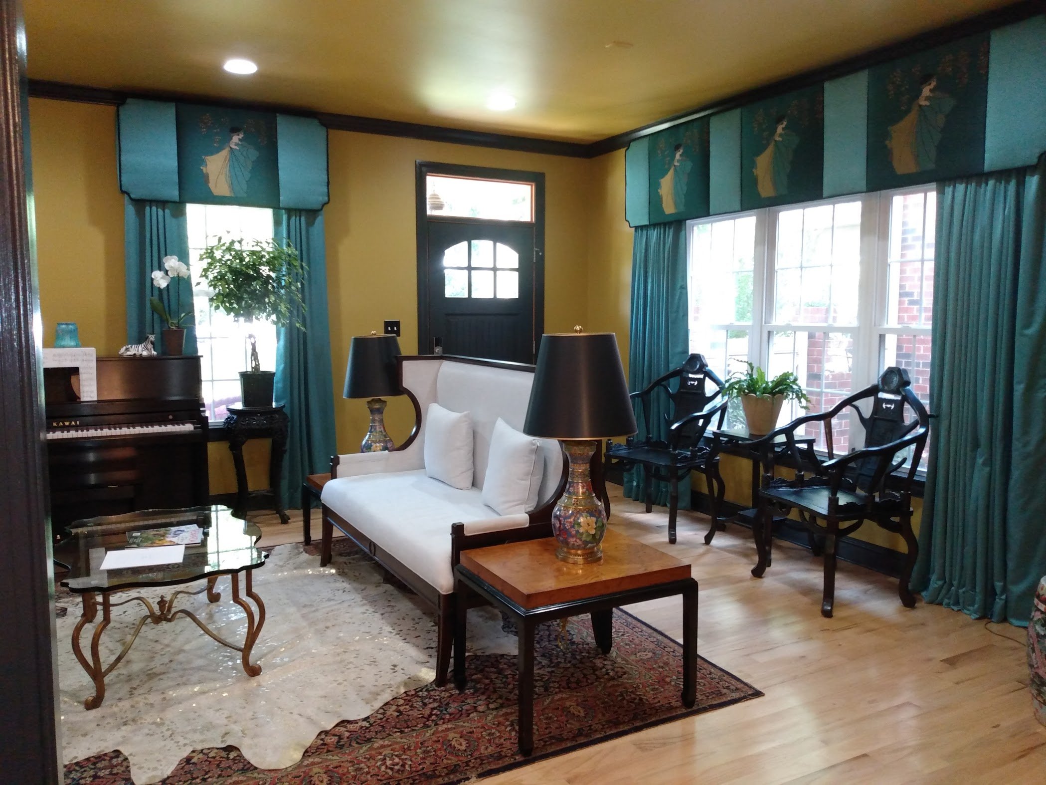 A cozy living room with yellow walls and large windows covered with turquoise curtains featuring a woman in traditional attire. The room has a white sofa with pillows, a wooden coffee table with decorative lamps, a piano with a potted orchid, and various chairs and rugs.