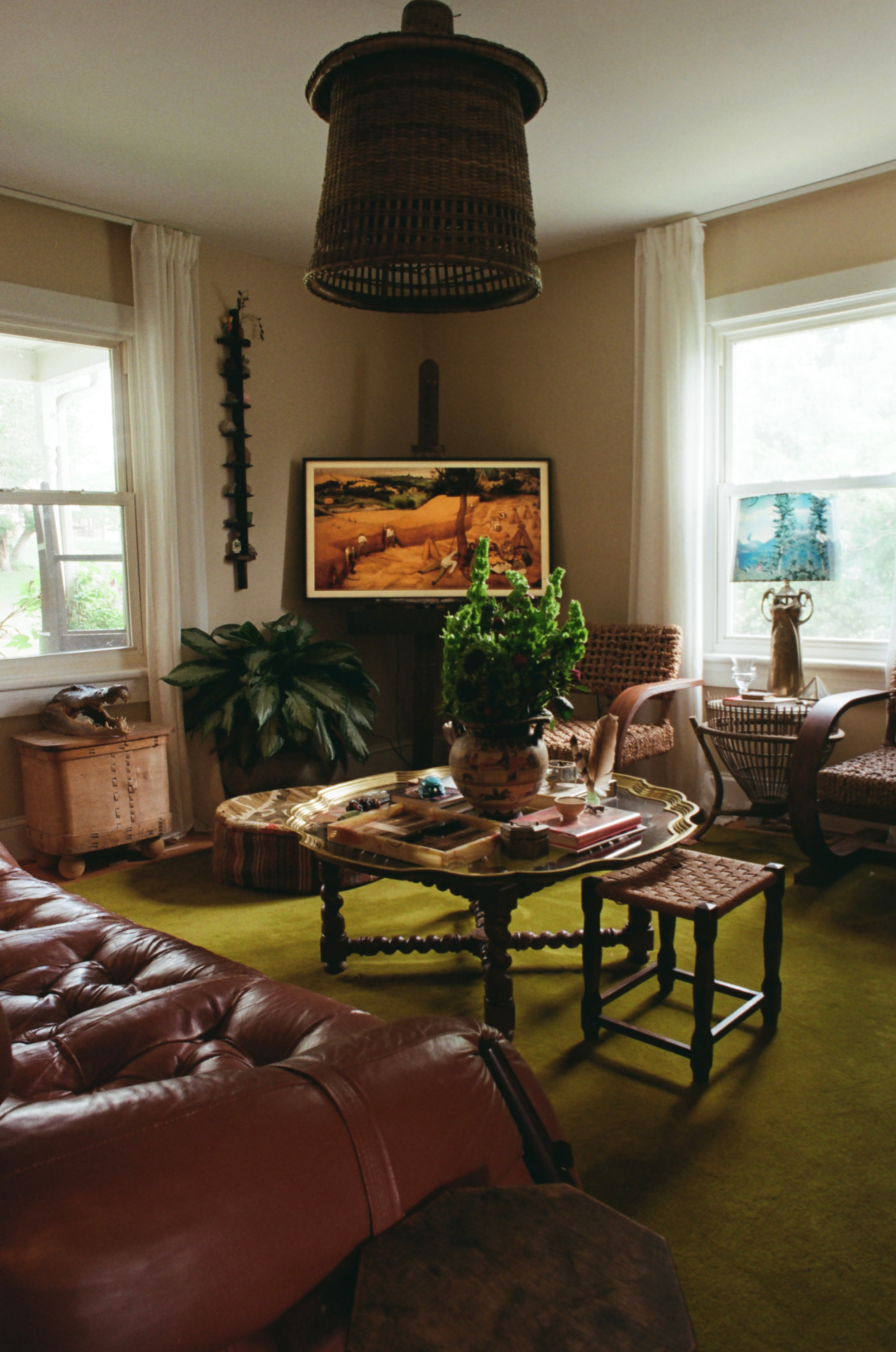 A cozy living room with a green carpet, a red leather couch, a wooden coffee table with a vase of flowers, large windows with white curtains, framed pictures, plants, and wicker chairs.