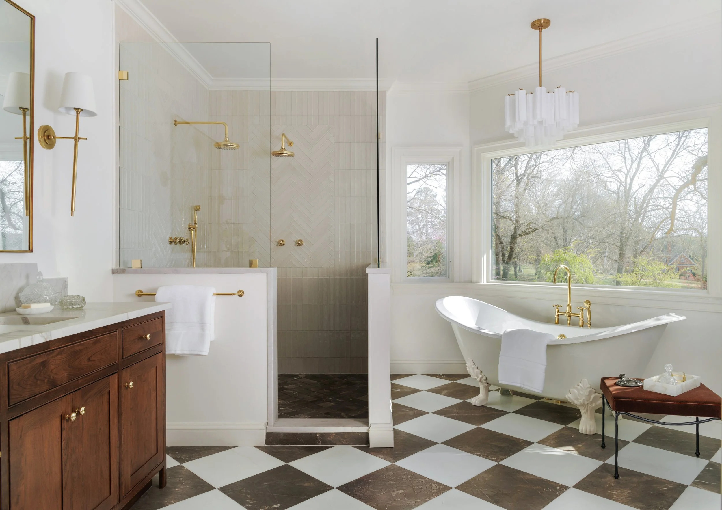 A spacious bathroom featuring a large window overlooking trees, a white clawfoot bathtub with gold fixtures, a wooden vanity with a marble countertop, a glass-enclosed shower with gold fixtures, checkered floor tiles, and elegant lighting fixtures, including a chandelier and wall sconces.