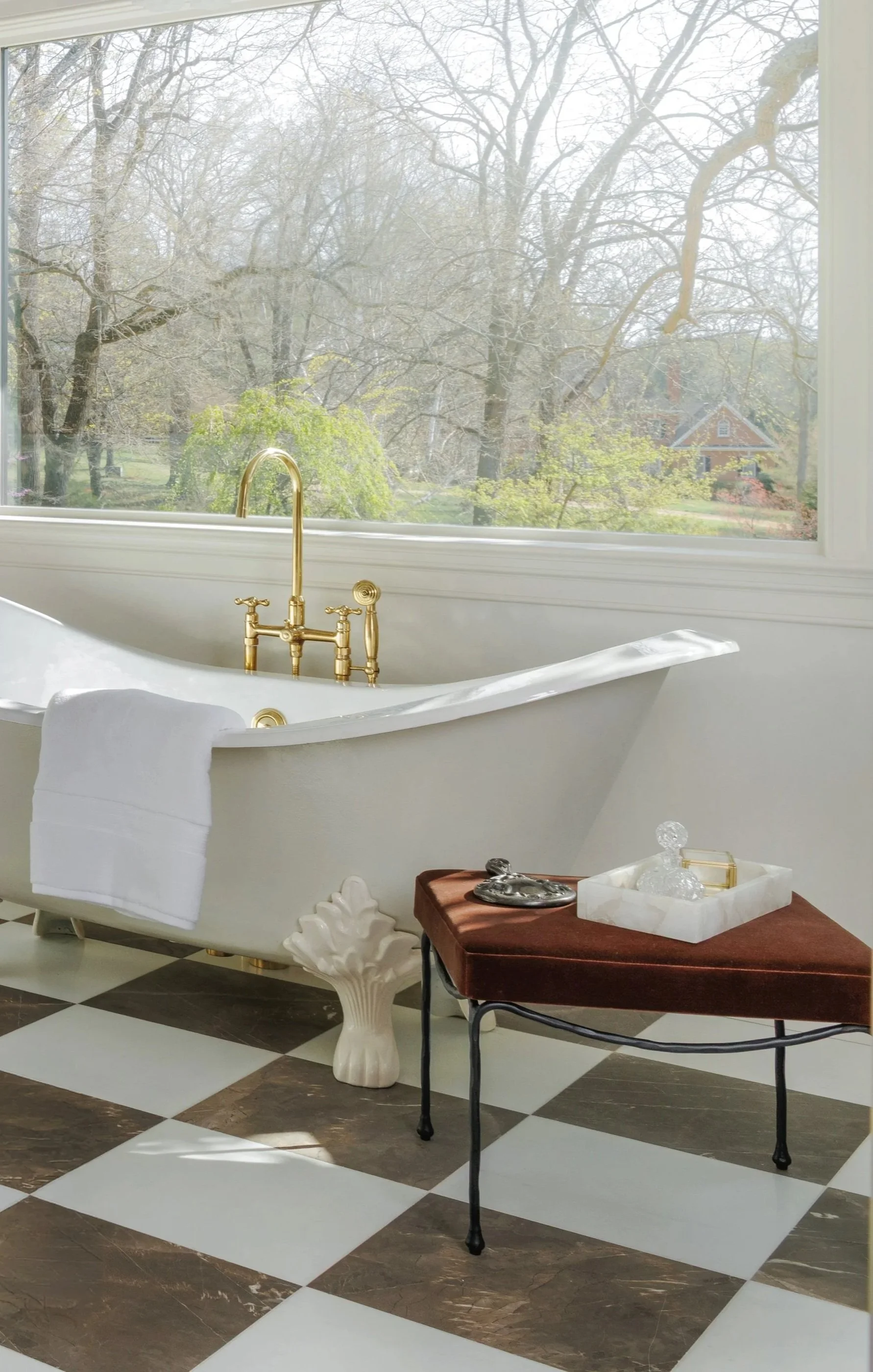 A white clawfoot bathtub with gold fixtures sits near a large window overlooking a garden with leafless trees and a house in the distance. Towel hangs over the edge of the bathtub. A wooden side table with a white marble tray holding glass and metallic objects is beside the tub, set on a checkered black-and-white marble floor.