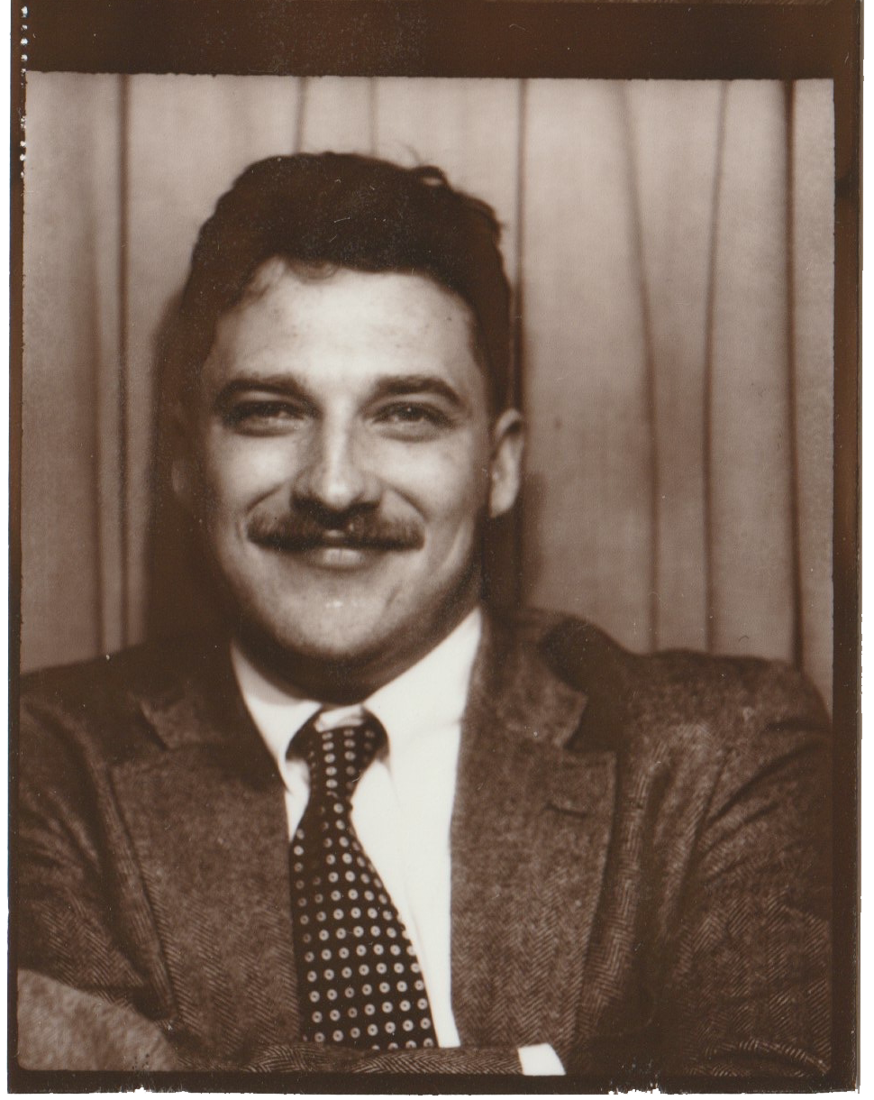 Black and white portrait of a man with dark hair, a mustache, and wearing a suit with a tie, smiling, against a striped backdrop.
