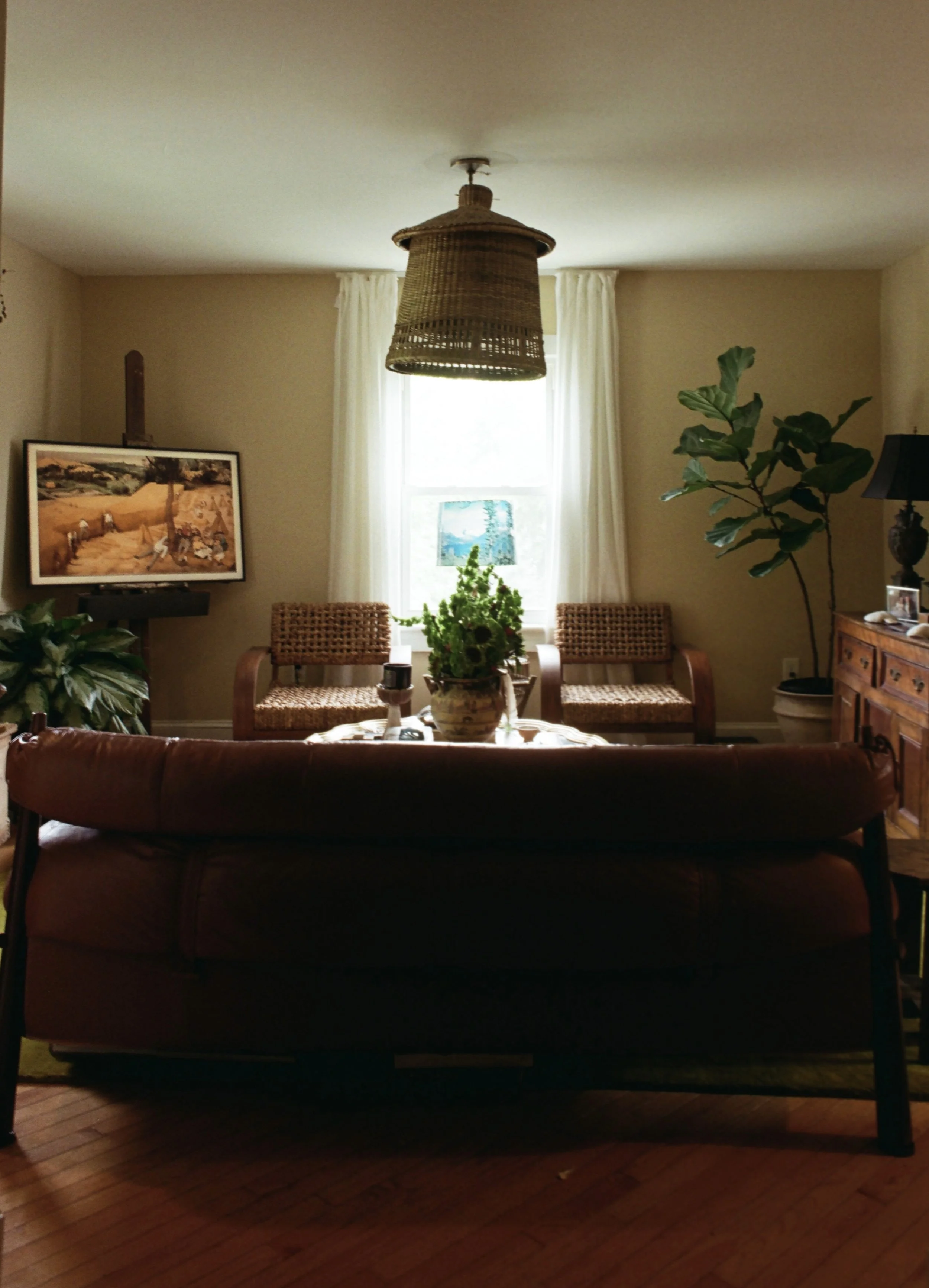 Living room with a brown leather sofa, a wooden dining table with rattan chairs, large window with white curtains, potted plants, framed artwork, and wicker ceiling light fixture.