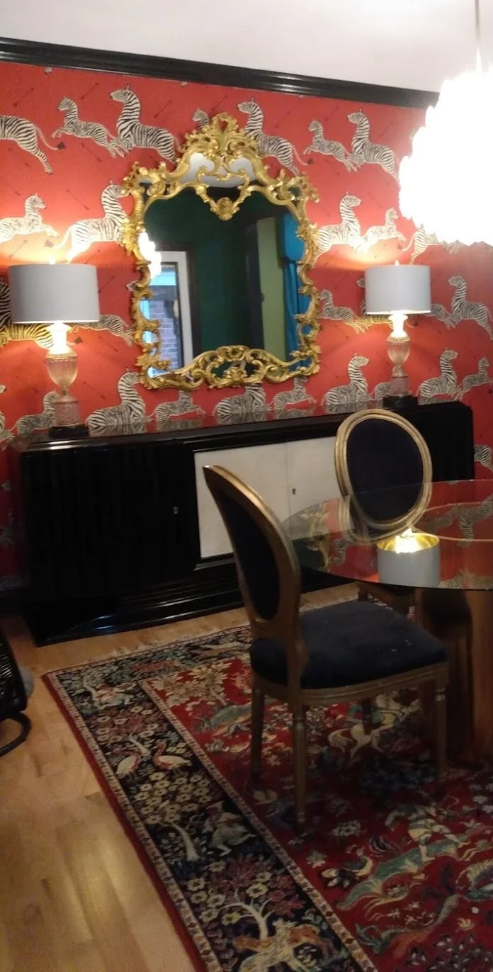 A dining room with red wallpaper featuring zebra patterns, an ornate gold-framed mirror, and matching lamps on a black sideboard. There is a round glass-top table with black and gold chairs, a patterned rug on the wooden floor, and a white ceiling light fixture.