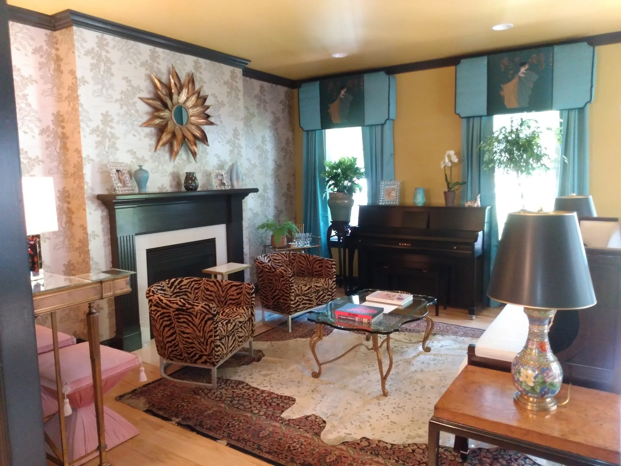 Living room with yellow walls, blue curtains, decorative items on a mantel, a black piano, a glass coffee table with books, patterned chairs, a pink windmill decorative piece, and a colorful vase.