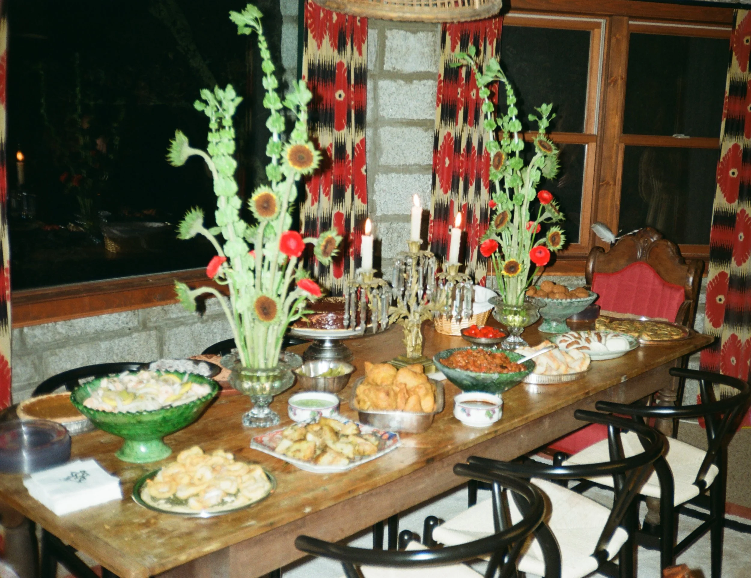 A dining table set for a celebration with various dishes, floral decorations, and candles in a room with patterned curtains and wooden furniture.