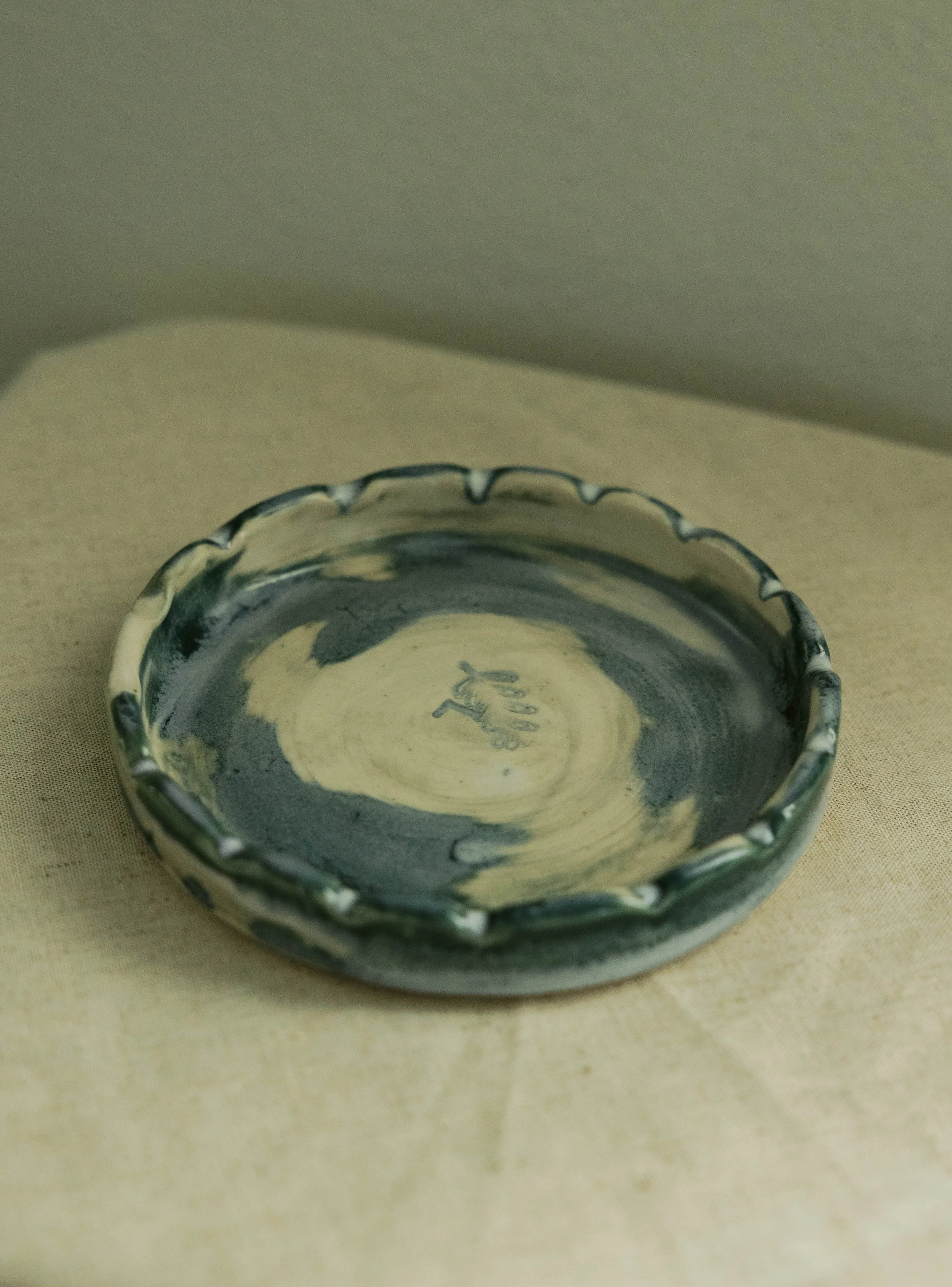 A small ceramic bowl with a scalloped edge, glazed in black and cream, resting on a beige fabric surface.