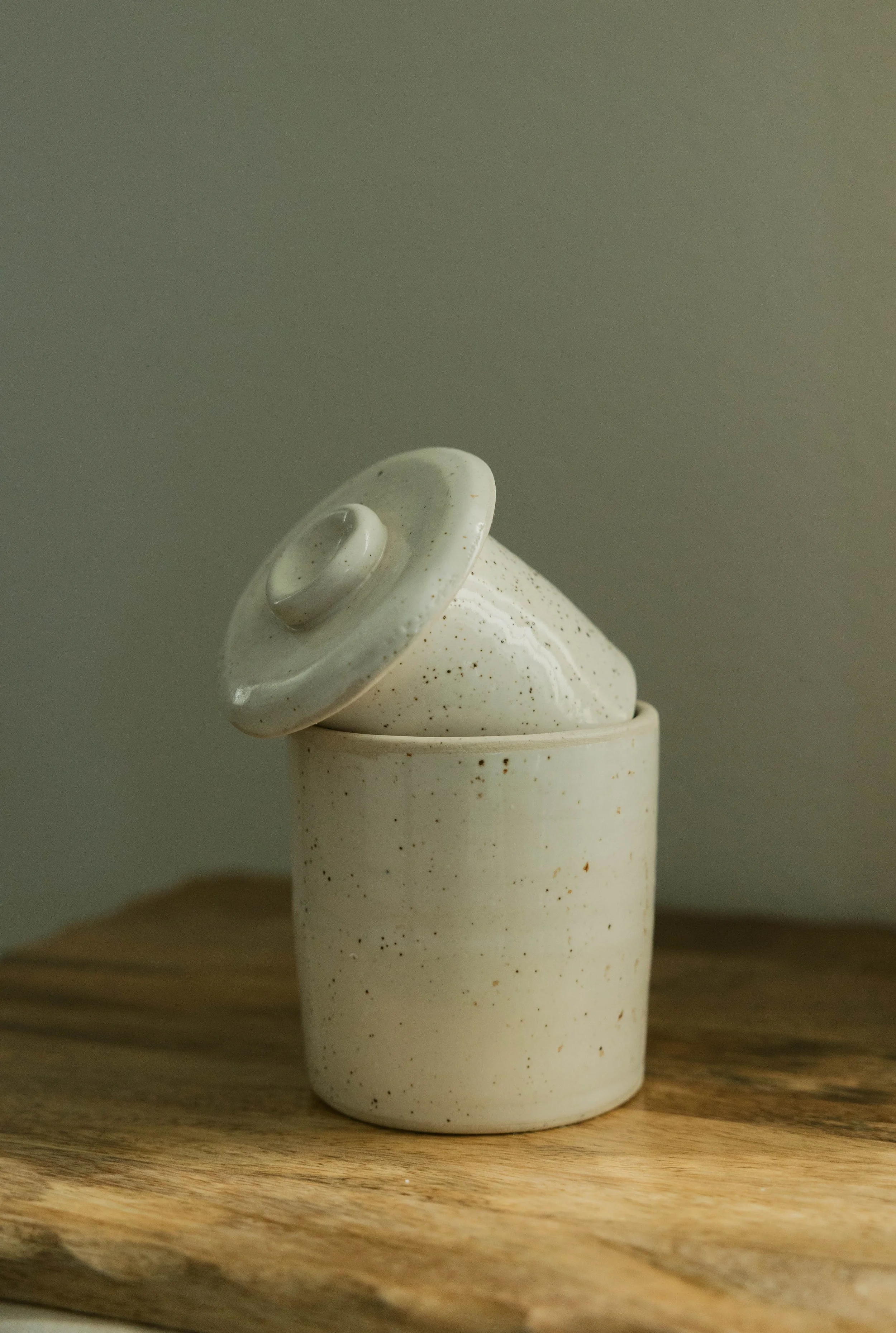 A white ceramic container with a removable lid, speckled with brown dots, placed on a wooden surface against a plain grey background.
