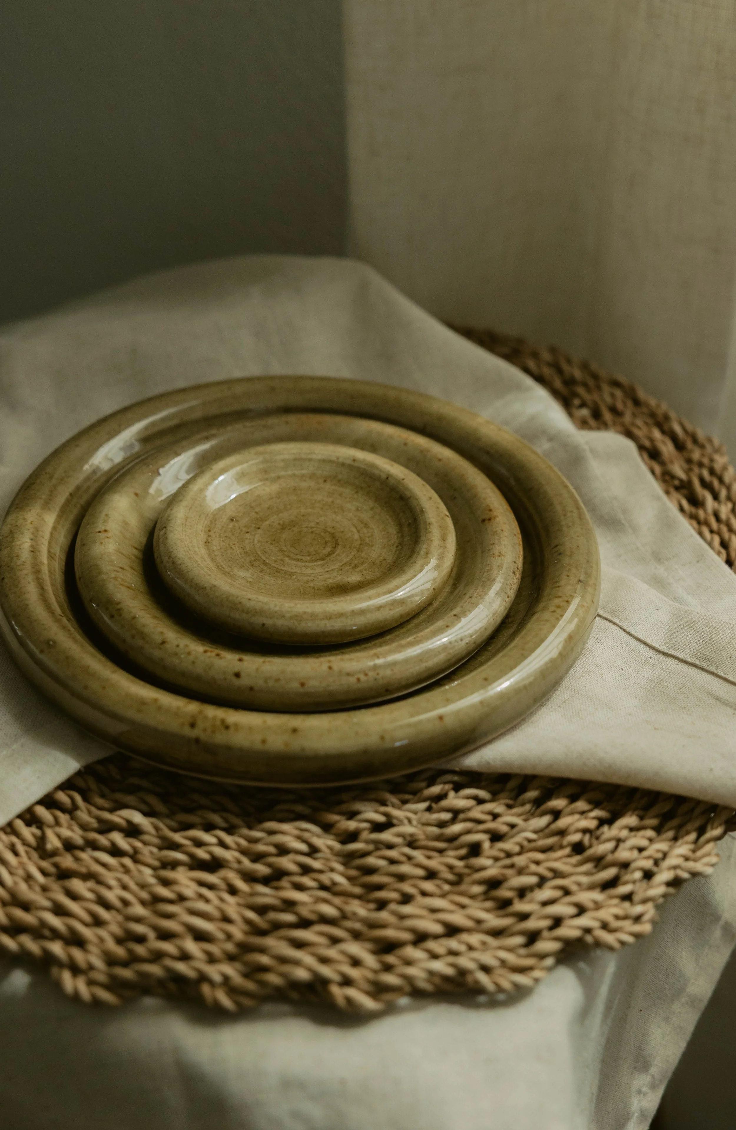 A beige ceramic plate with a concentric circular design, placed on a white cloth, with a woven rattan tray underneath and a beige fabric background.