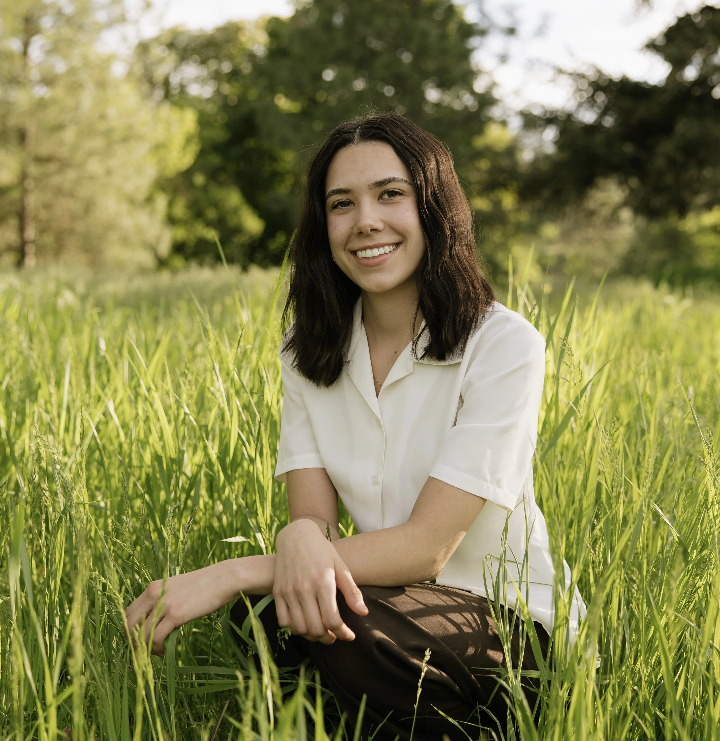 Young woman sitting in a green field with tall grass, smiling, with trees in the background during daylight.