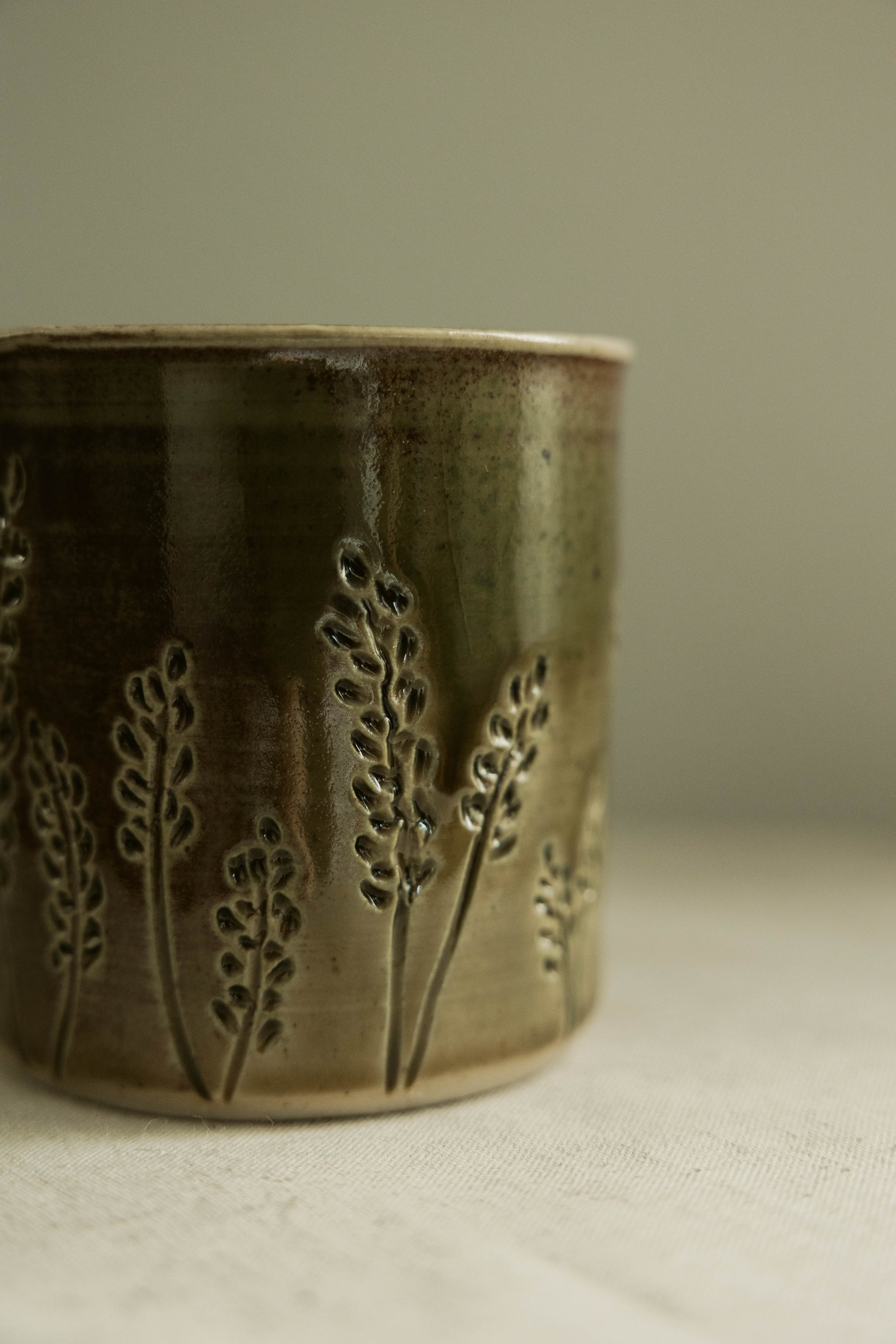 Close-up photograph of a ceramic cup with a textured pattern of small, raised floral designs, made from a brownish-green glazed ceramic material.