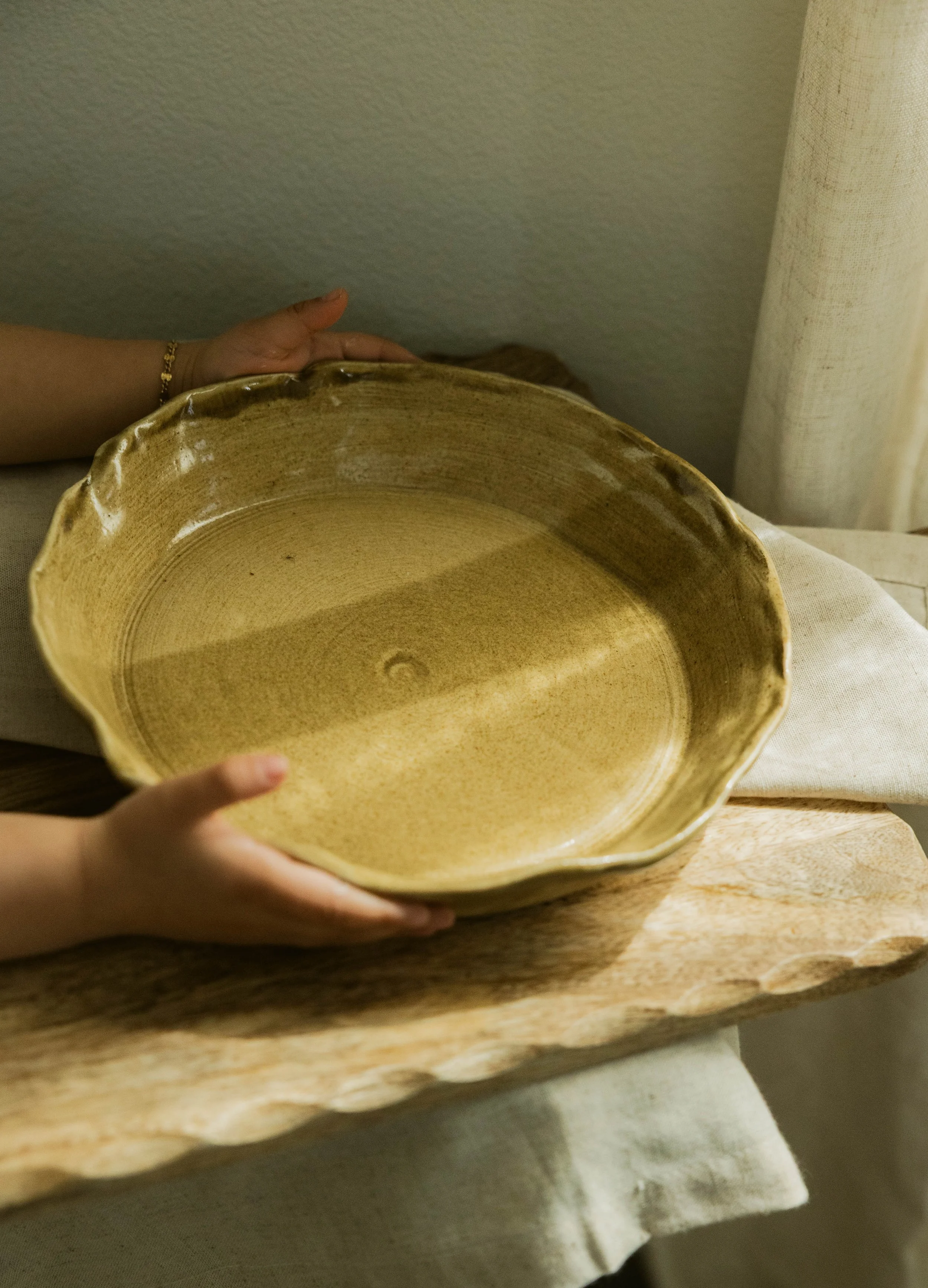 A person holding a large, beige, ceramic bowl with both hands, standing over a wooden table covered with a white cloth, with a beige curtain in the background.