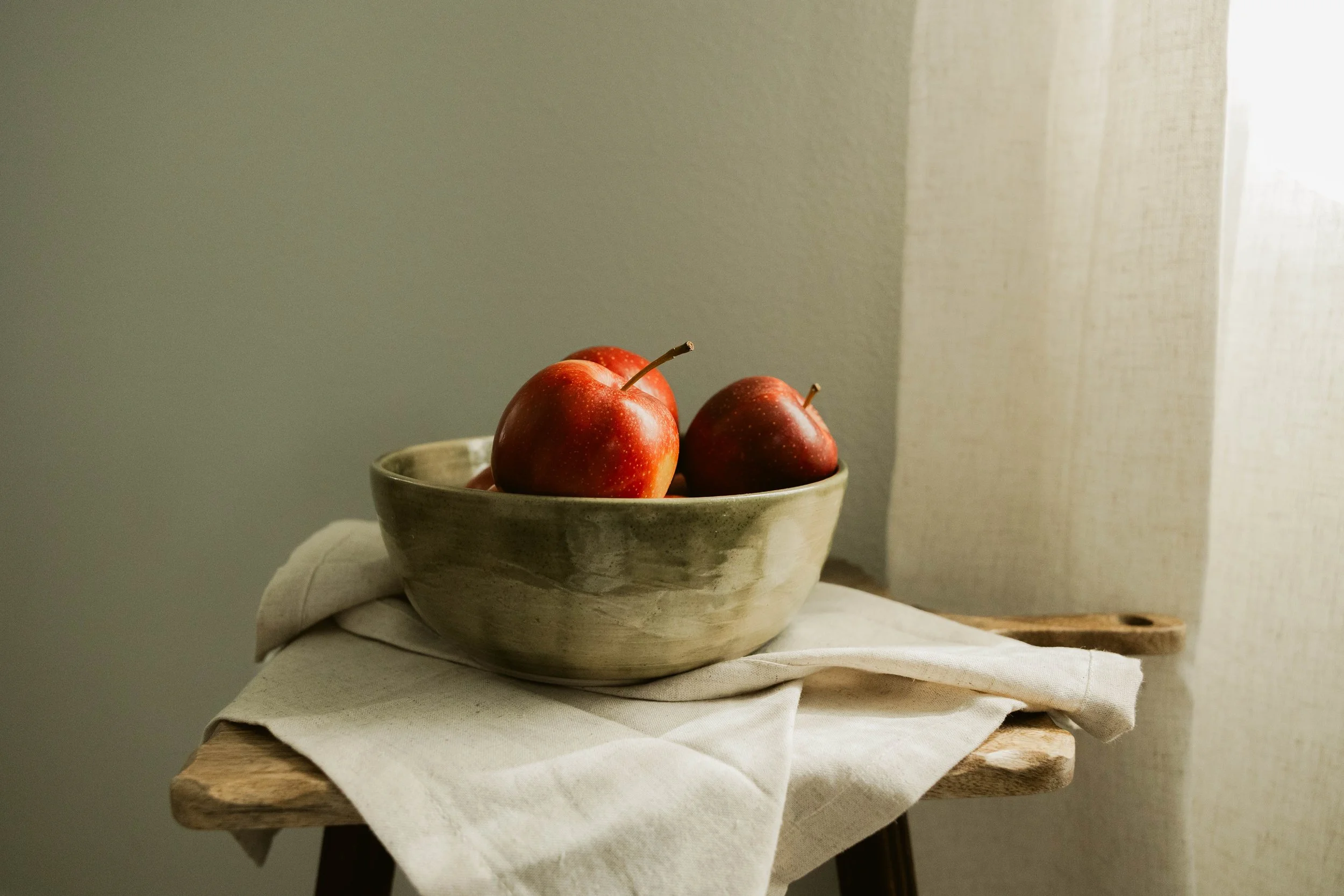 Three red apples in a ceramic bowl on a white cloth-covered wooden table near a light-colored curtain.
