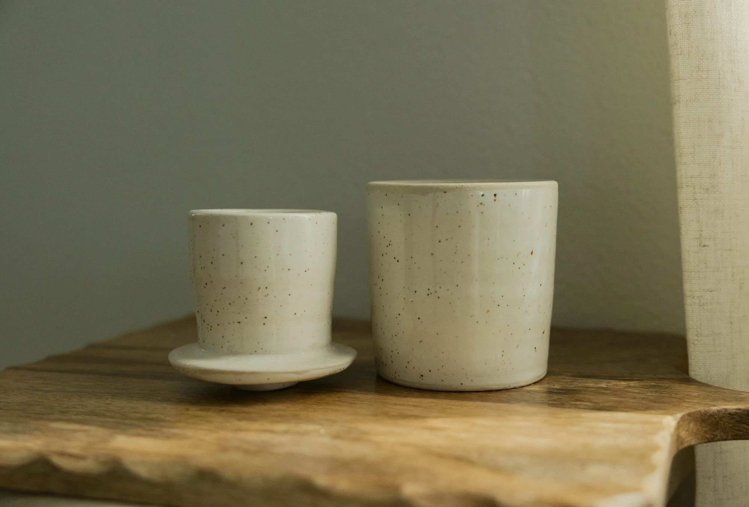 Two speckled white ceramic cups, one with a matching saucer, placed on a wooden surface against a plain wall background.