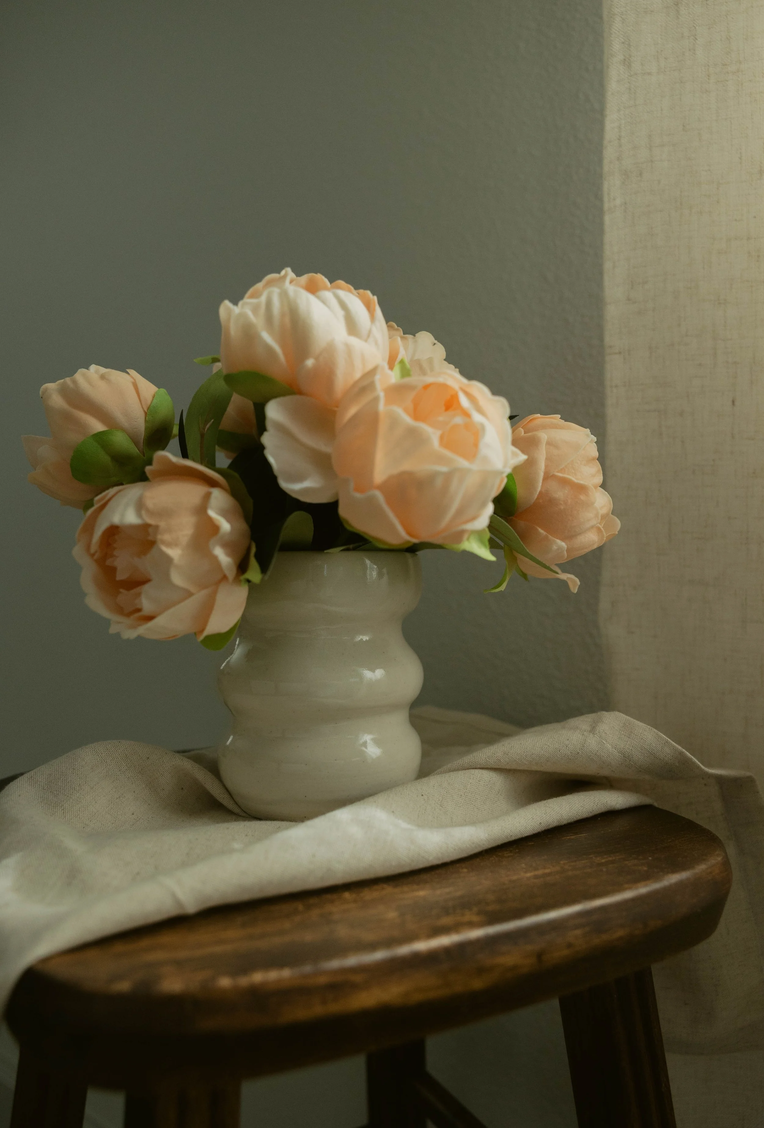 A white ceramic vase with peach-colored roses, placed on a wooden tray with a beige cloth underneath, against a neutral wall background.