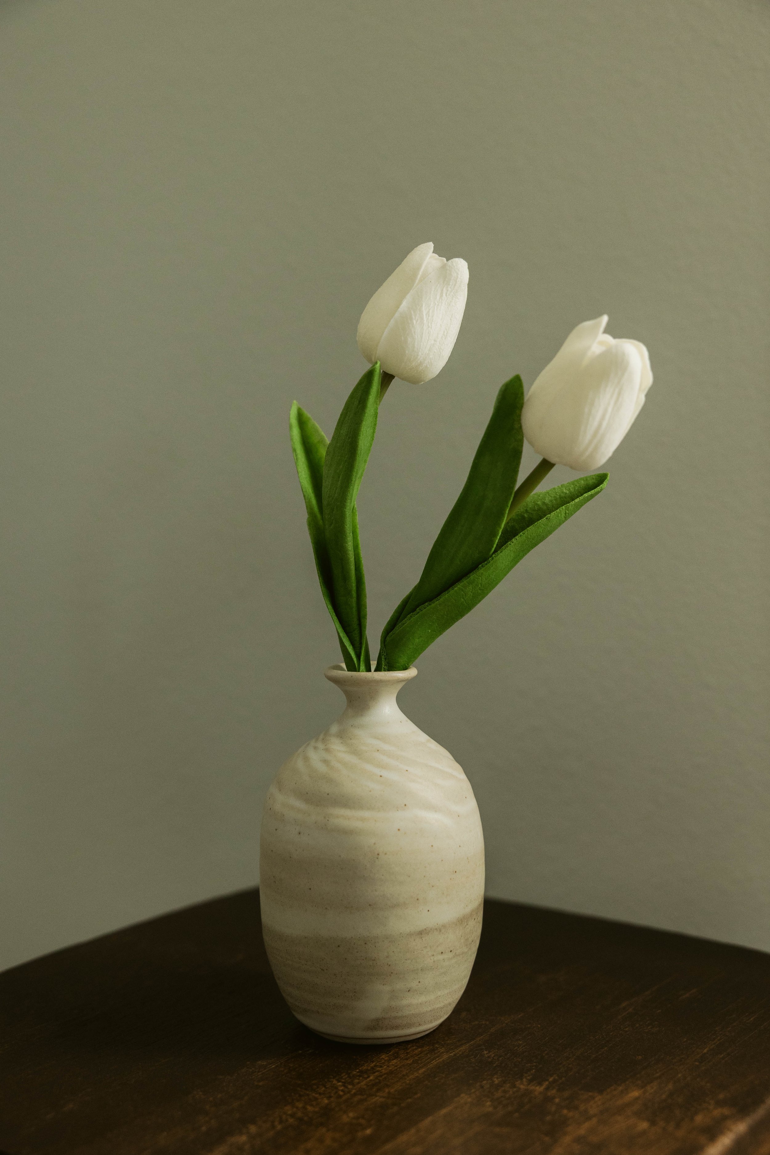 A white ceramic vase with subtle horizontal stripes holding two white tulips with green leaves, placed on a dark wooden surface against a plain gray background.