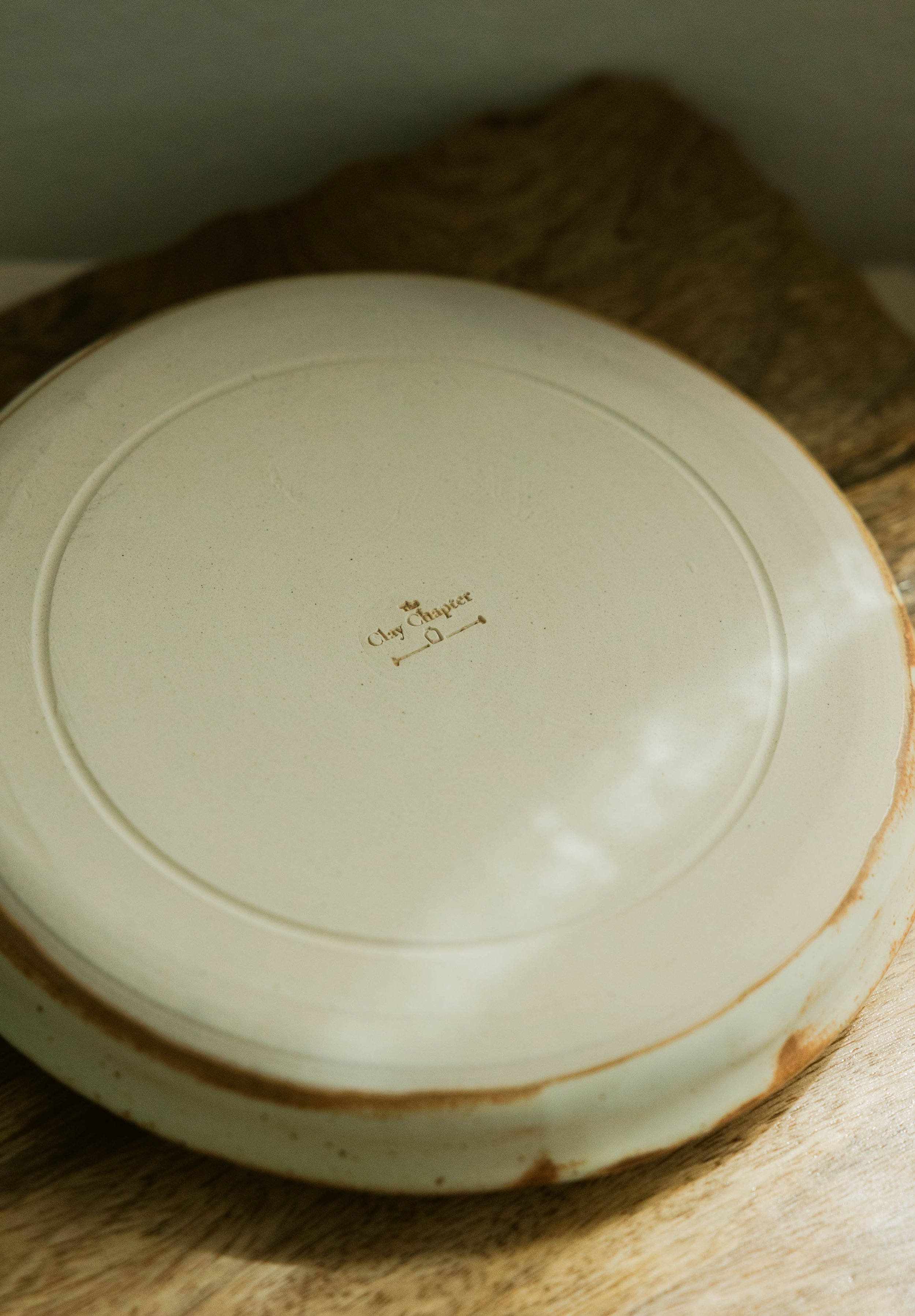 Close-up of the bottom of a white ceramic plate with a gold-colored marking that says 'Clay Chapter' and an illustration of a key, resting on a wooden surface.