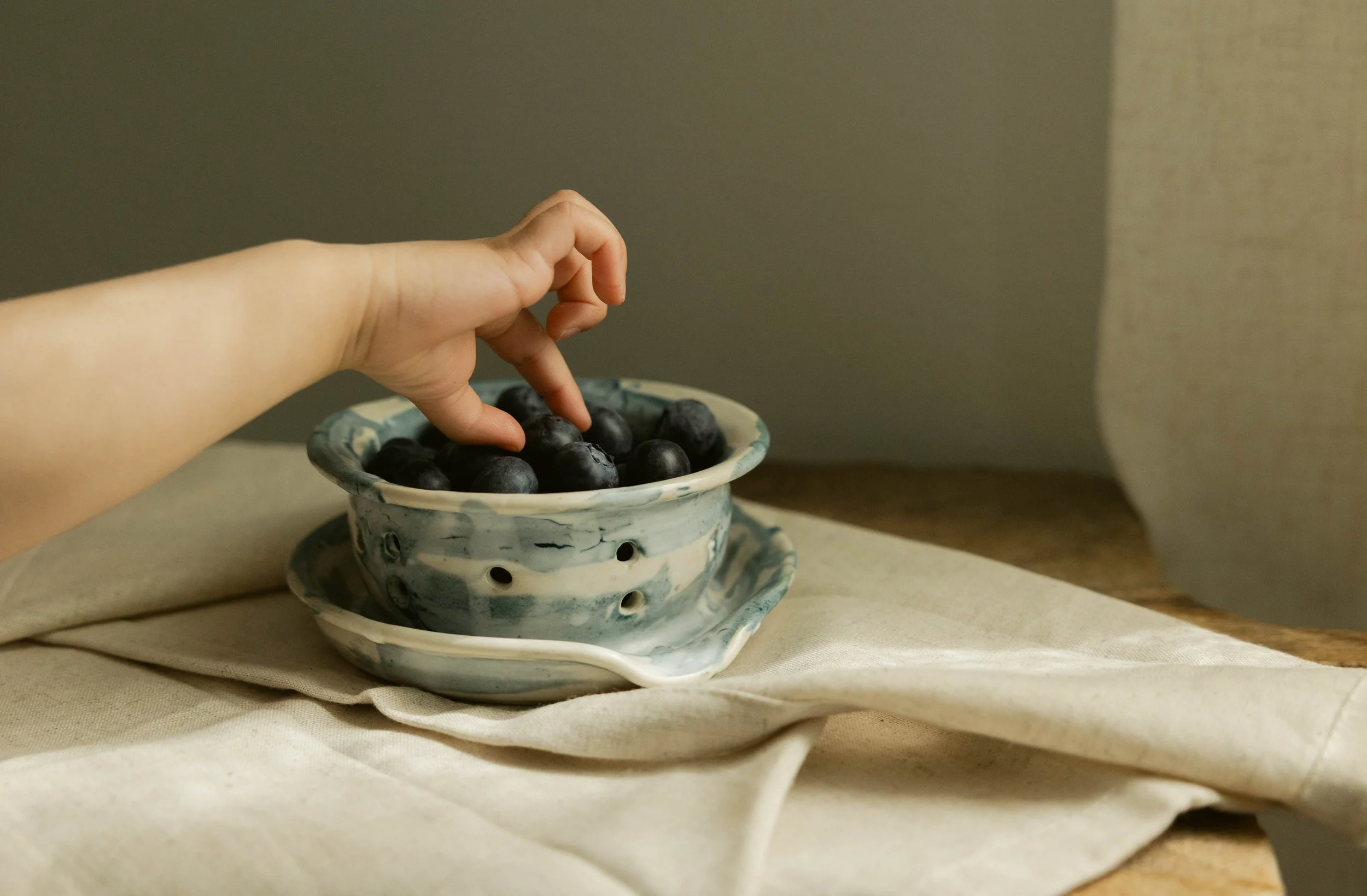 Hand reaching for blueberries in a ceramic bowl on a table.