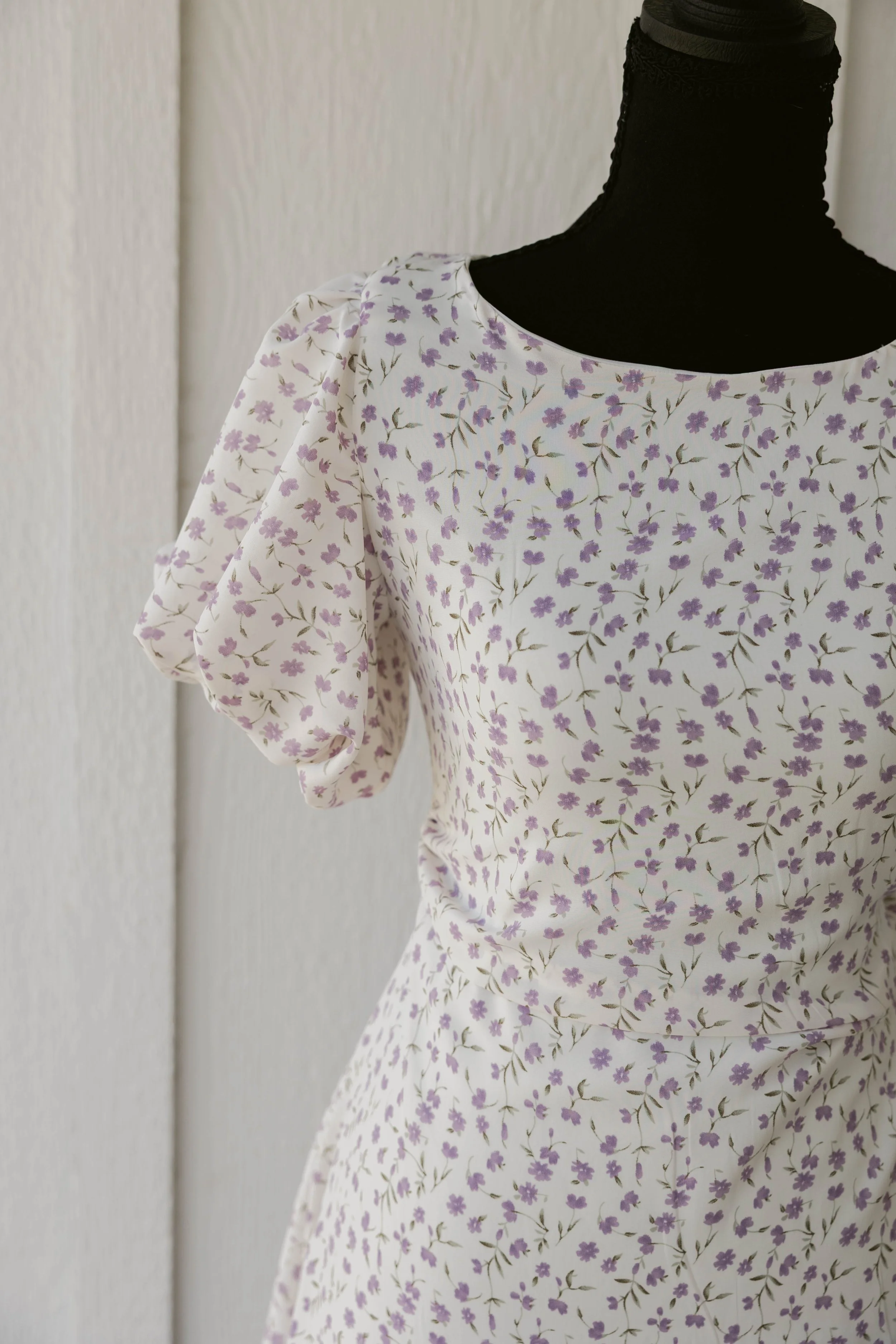 A mannequin dressed in a white dress with a purple floral pattern, set against a plain beige wall.