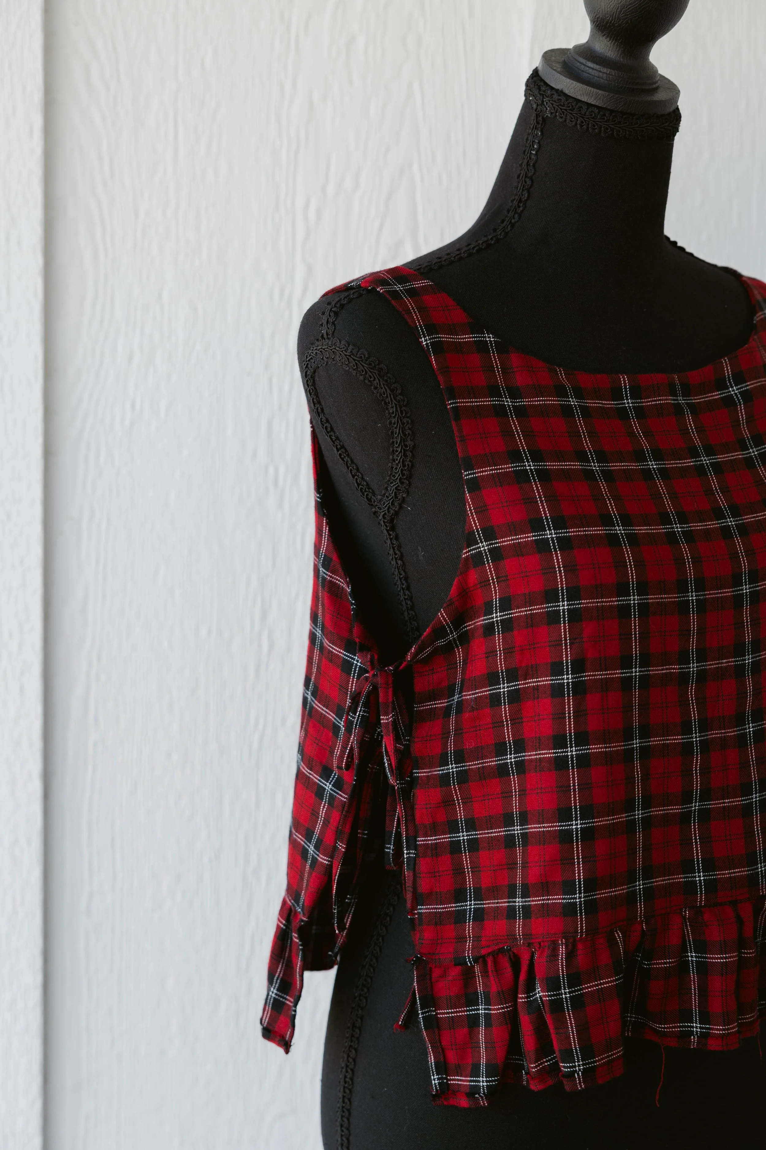 Black mannequin wearing a red and black plaid sleeveless top with tie detail on the side, against a plain white wall background.