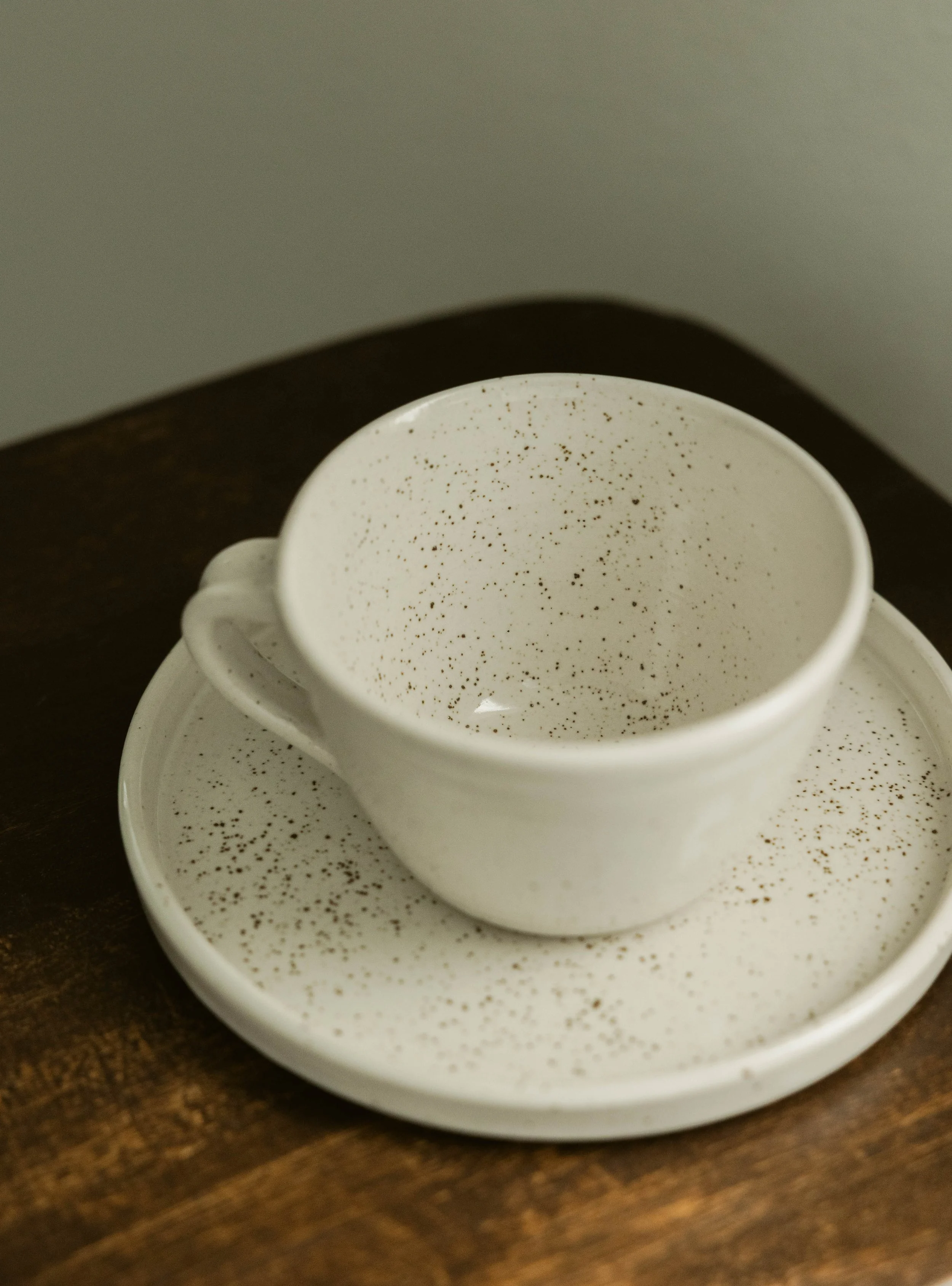 A white ceramic coffee cup with black speckles on a matching saucer, placed on a dark wooden surface.