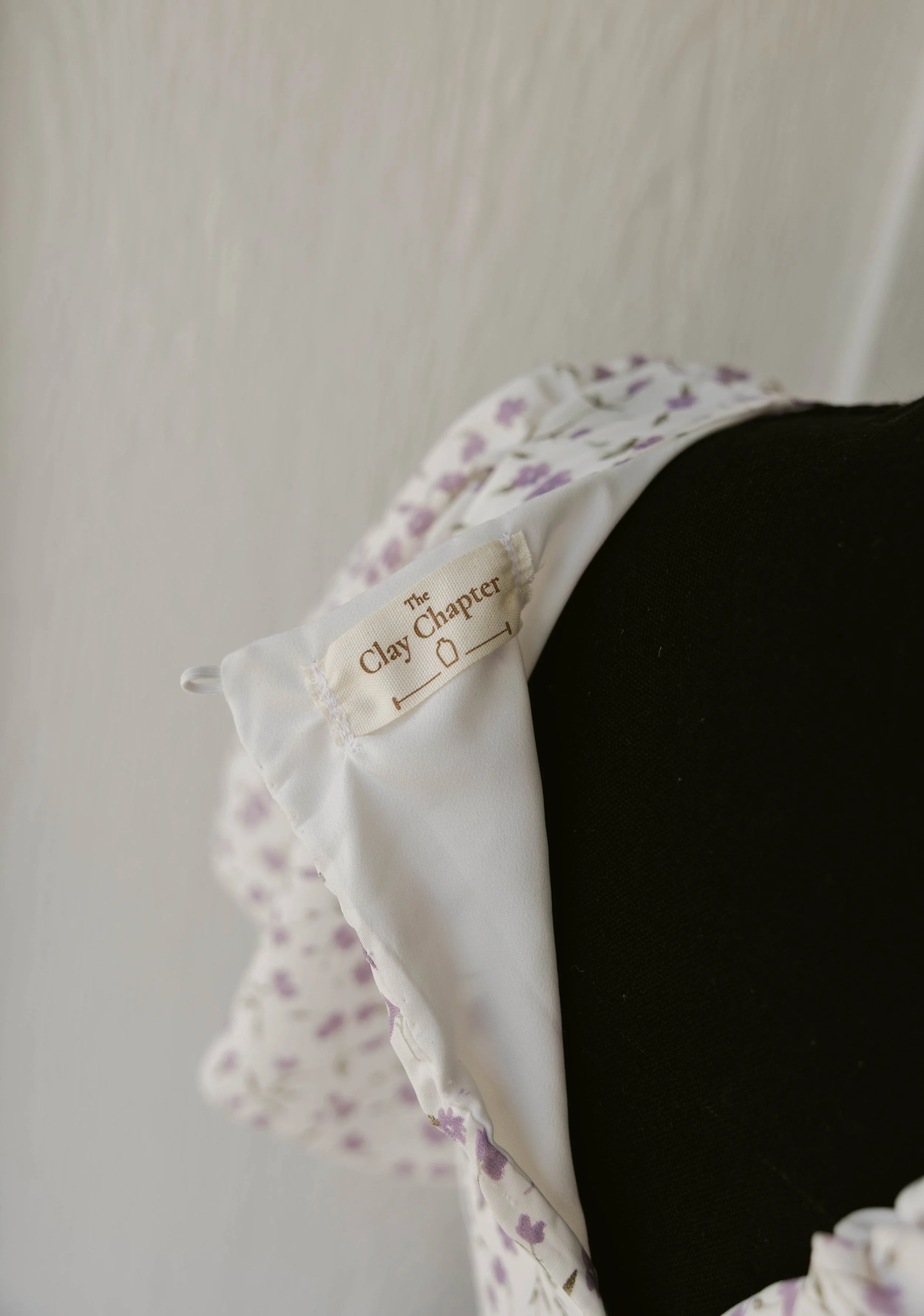 Close-up of a white fabric tag with beige embroidery reading 'The Clay Chapter' attached to a floral-patterned garment, displayed on a black mannequin against a plain background.