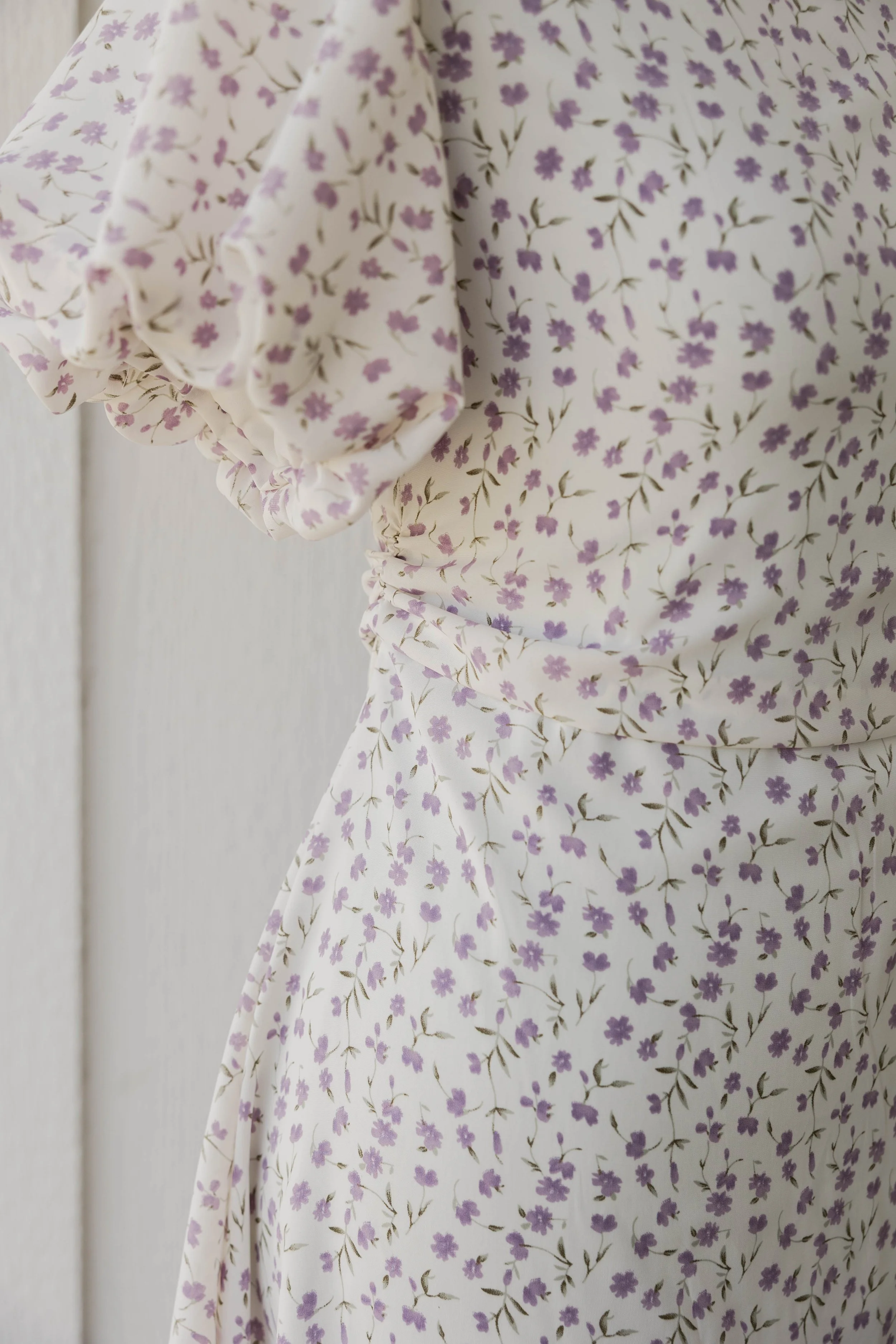 Close-up of a white fabric dress with a pattern of small purple flowers and green stems, featuring puffed sleeves with elastic at the cuffs.