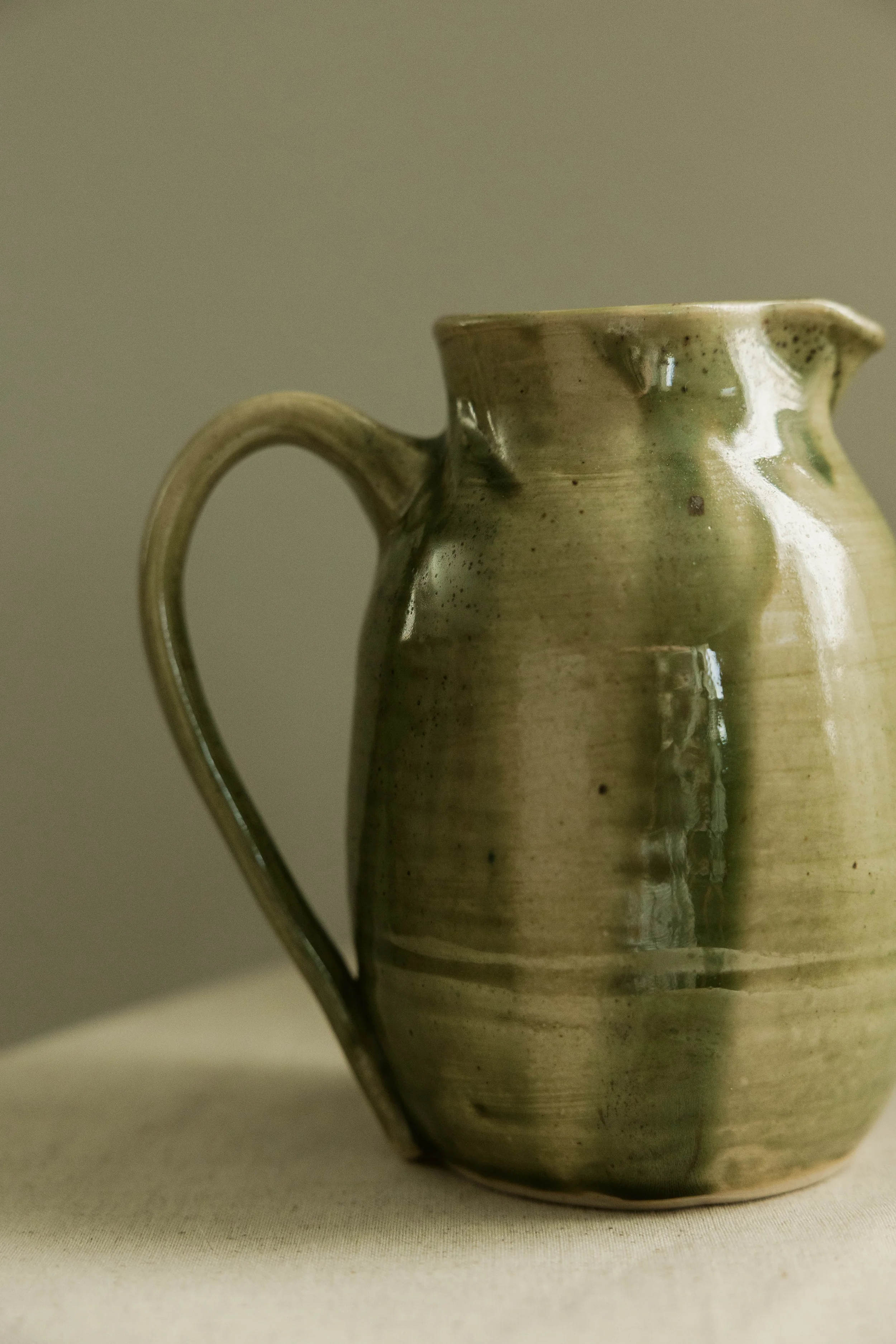 A ceramic pitcher with a handle, glazed in earthy green and brown tones, standing on a neutral surface.