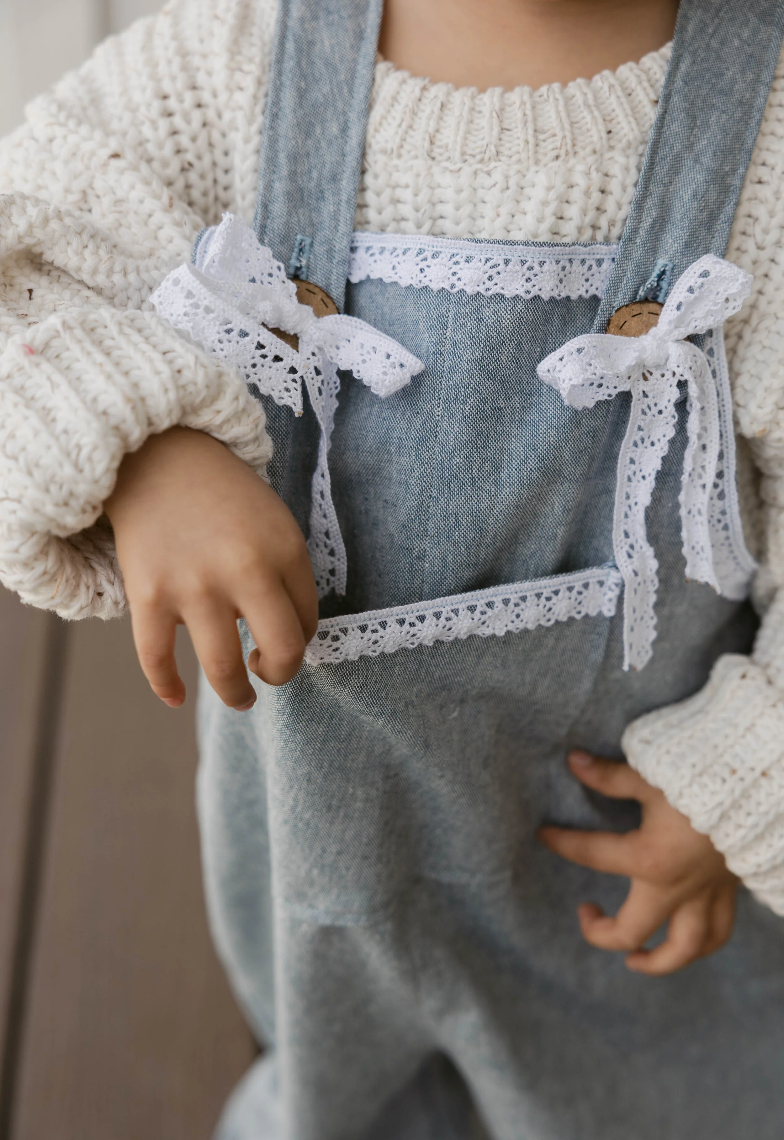 Close-up of a child's torso wearing a cream knit sweater and a denim apron with lace and tie details, and a hand resting near the waist.