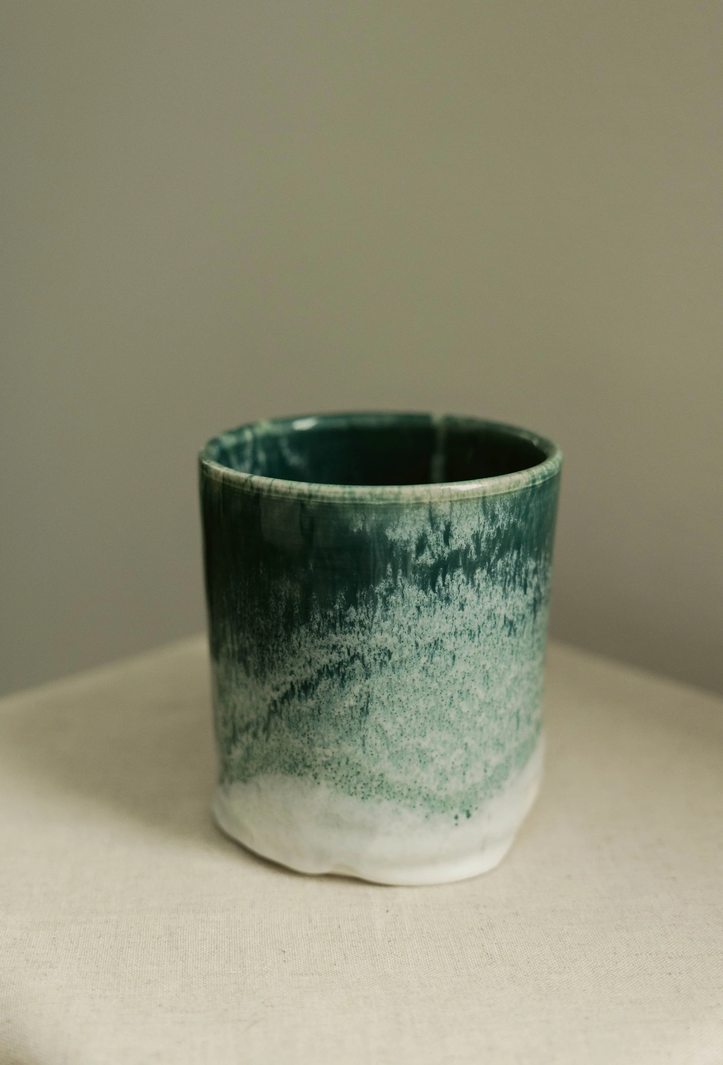 A ceramic cup with a gradient glaze from dark green at the top to white at the bottom, placed on a plain surface against a neutral background.