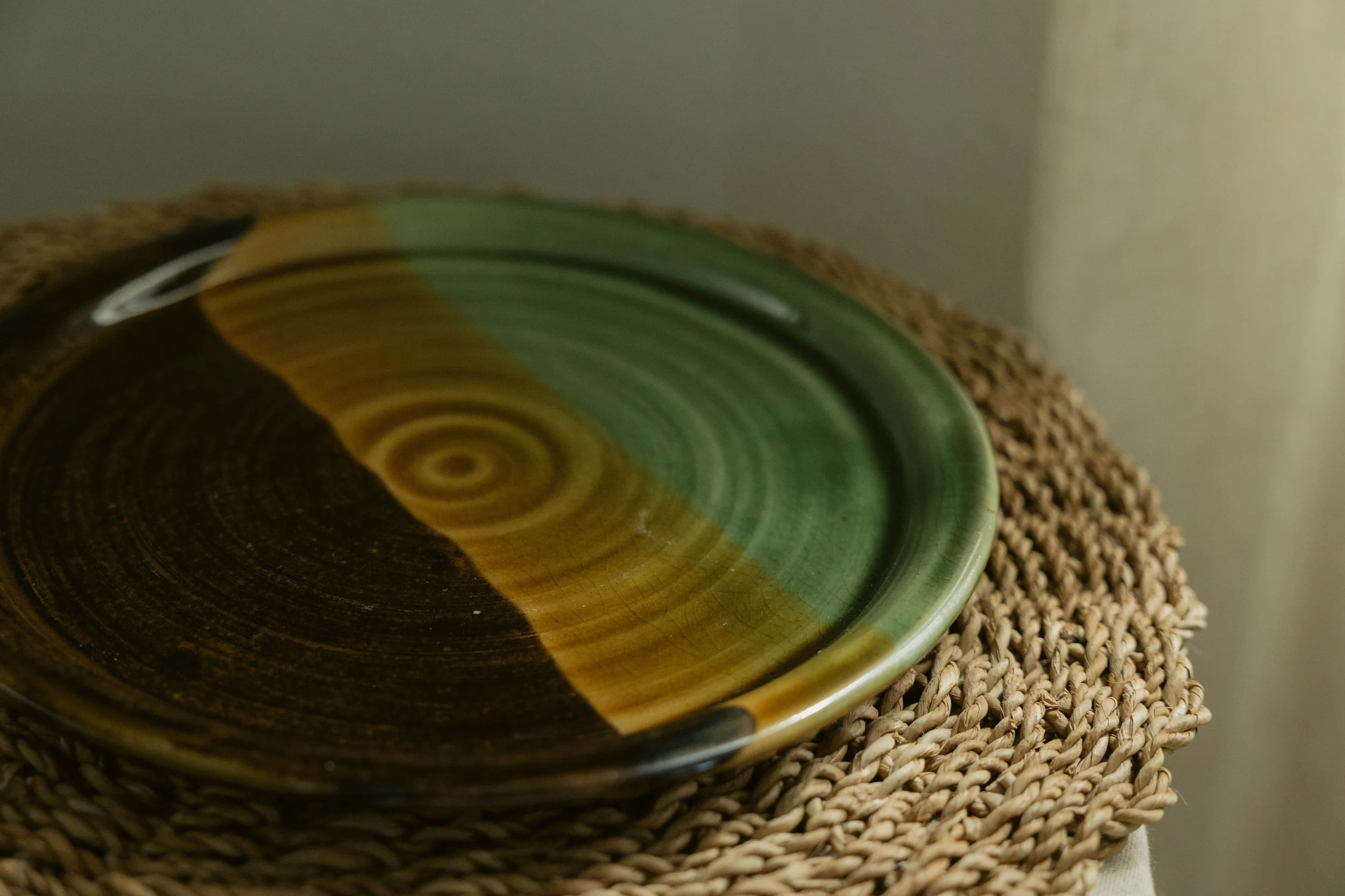 Close-up of a colorful pottery bowl with concentric rings of green, brown, and beige, resting on a woven mat.