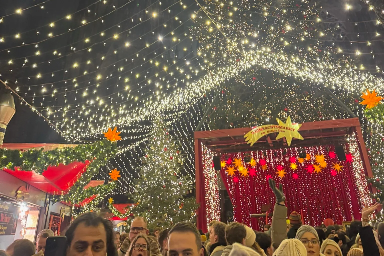A dense canopy of white fairy lights above a crowded evening Christmas market in Cologne, with lit stalls and star decorations below.