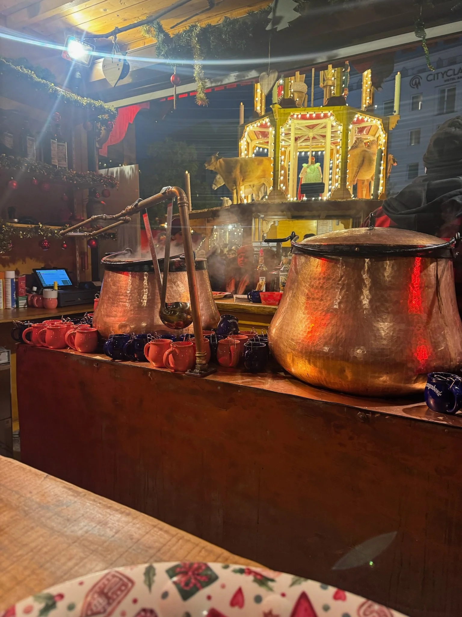 Steaming copper vats of mulled wine on a market stall counter, with rows of red and blue ceramic mugs and a lit carousel decoration behind.