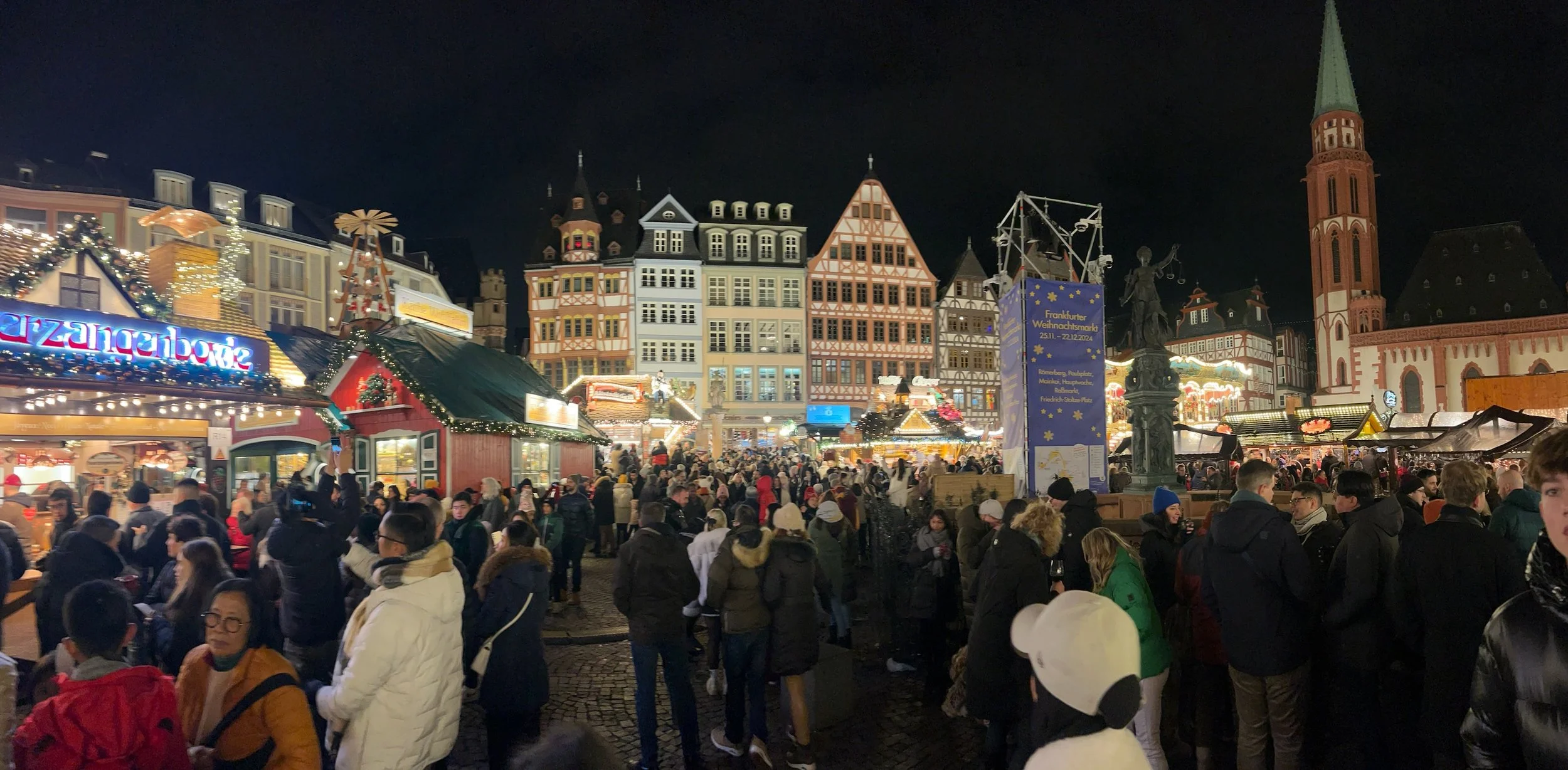 Crowds in winter coats at a night-time Christmas market in Frankfurt's Romerberg square, with lit wooden stalls, half-timbered houses and a church tower behind.