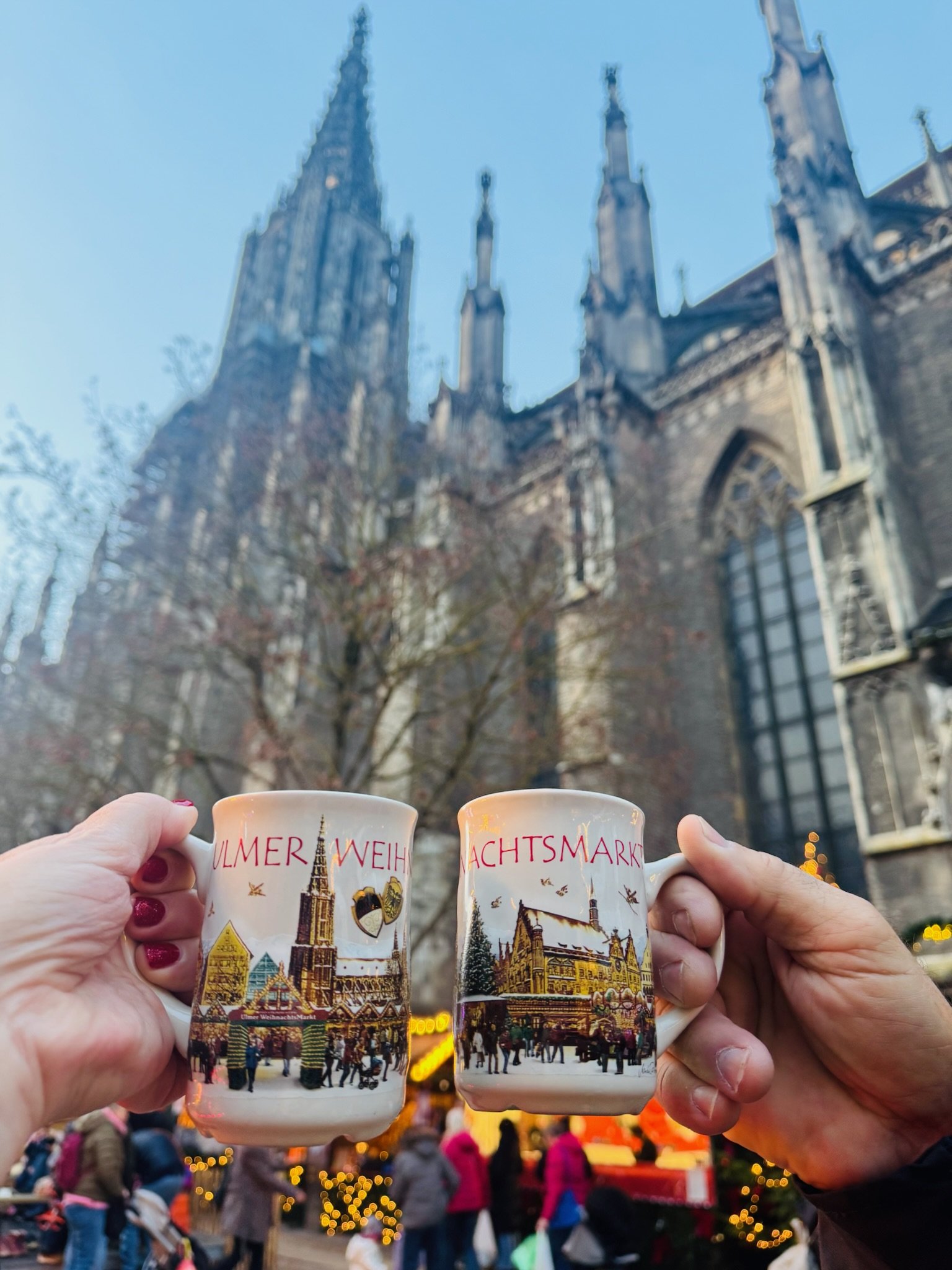 Two hands holding up decorated Ulm Christmas market mugs in front of a Gothic church, with market stalls and shoppers below.