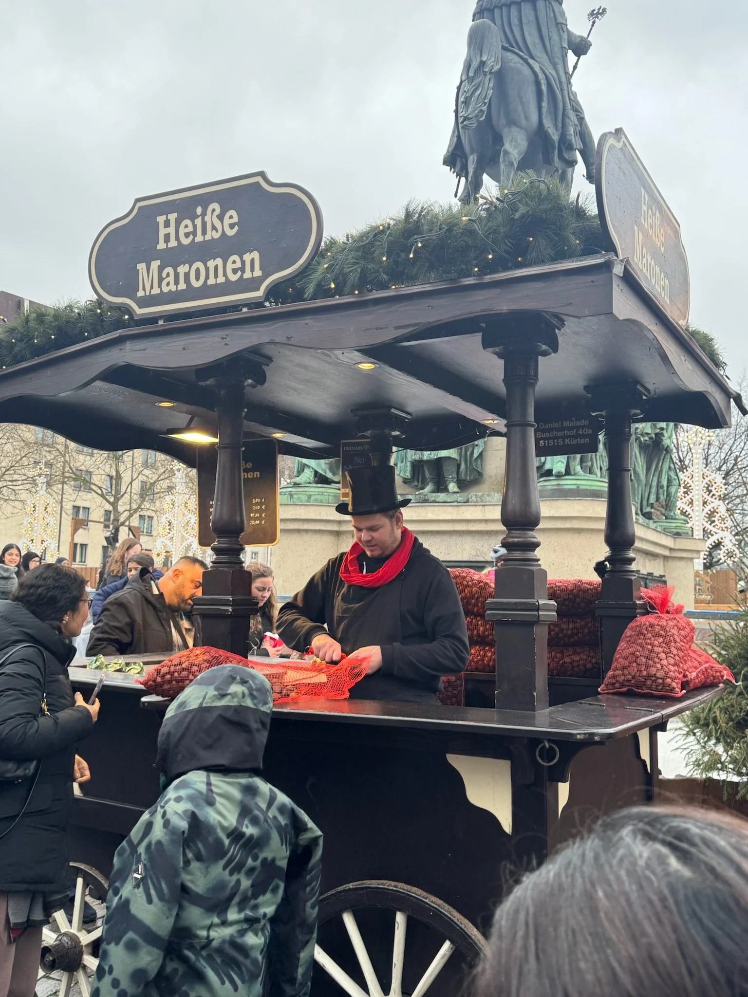 A vendor in a black coat and top hat serves customers from a wooden cart selling hot roasted chestnuts, with red net sacks of chestnuts alongside.