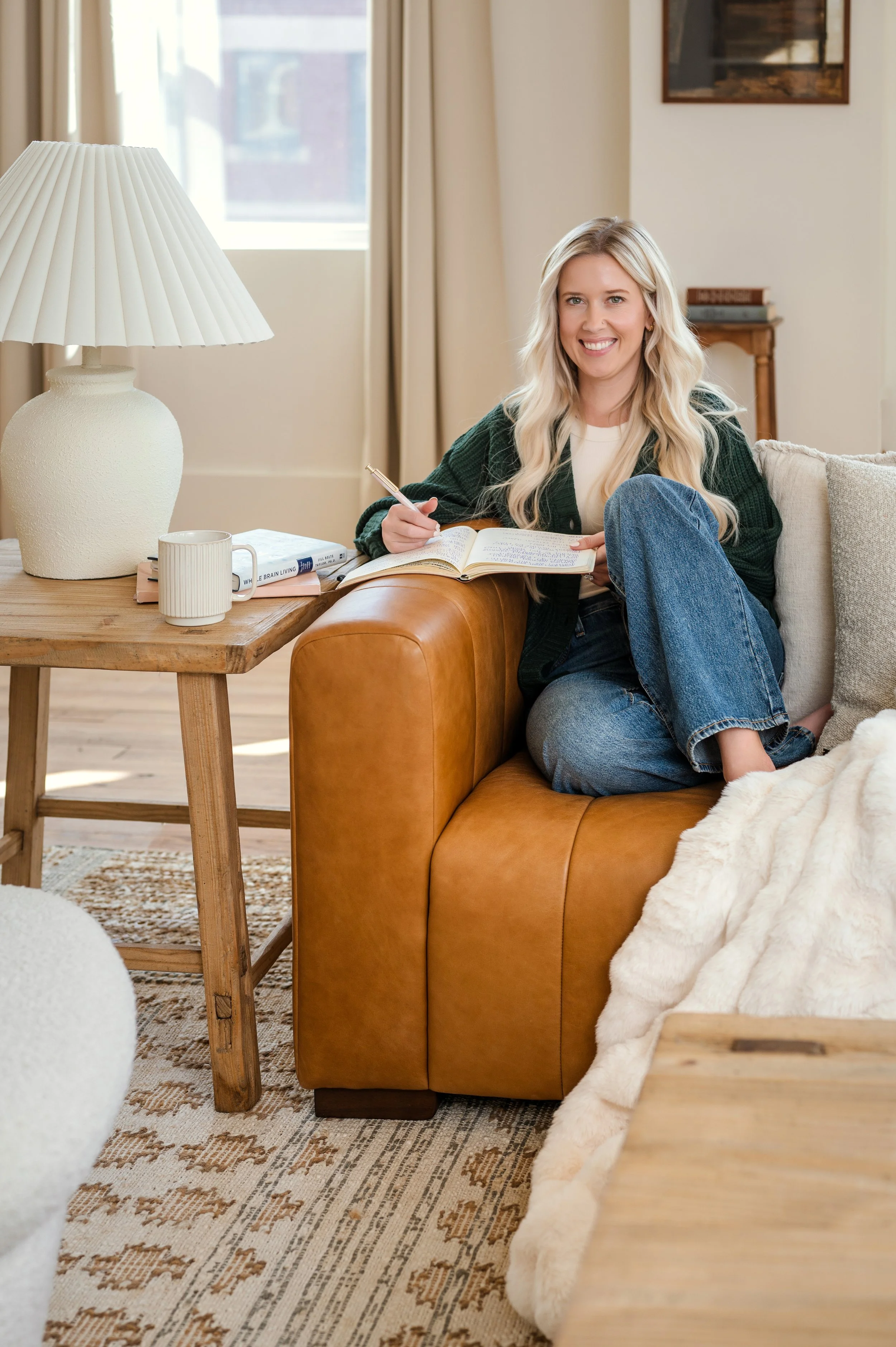 A woman with long blonde hair, sitting on a tan leather sofa, smiling, and writing in a notebook. She is wearing a white shirt, blue jeans, and a dark green cardigan. There's a wooden table with a white lamp, a mug, and books beside her, in a well-lit living room with beige walls and curtains.