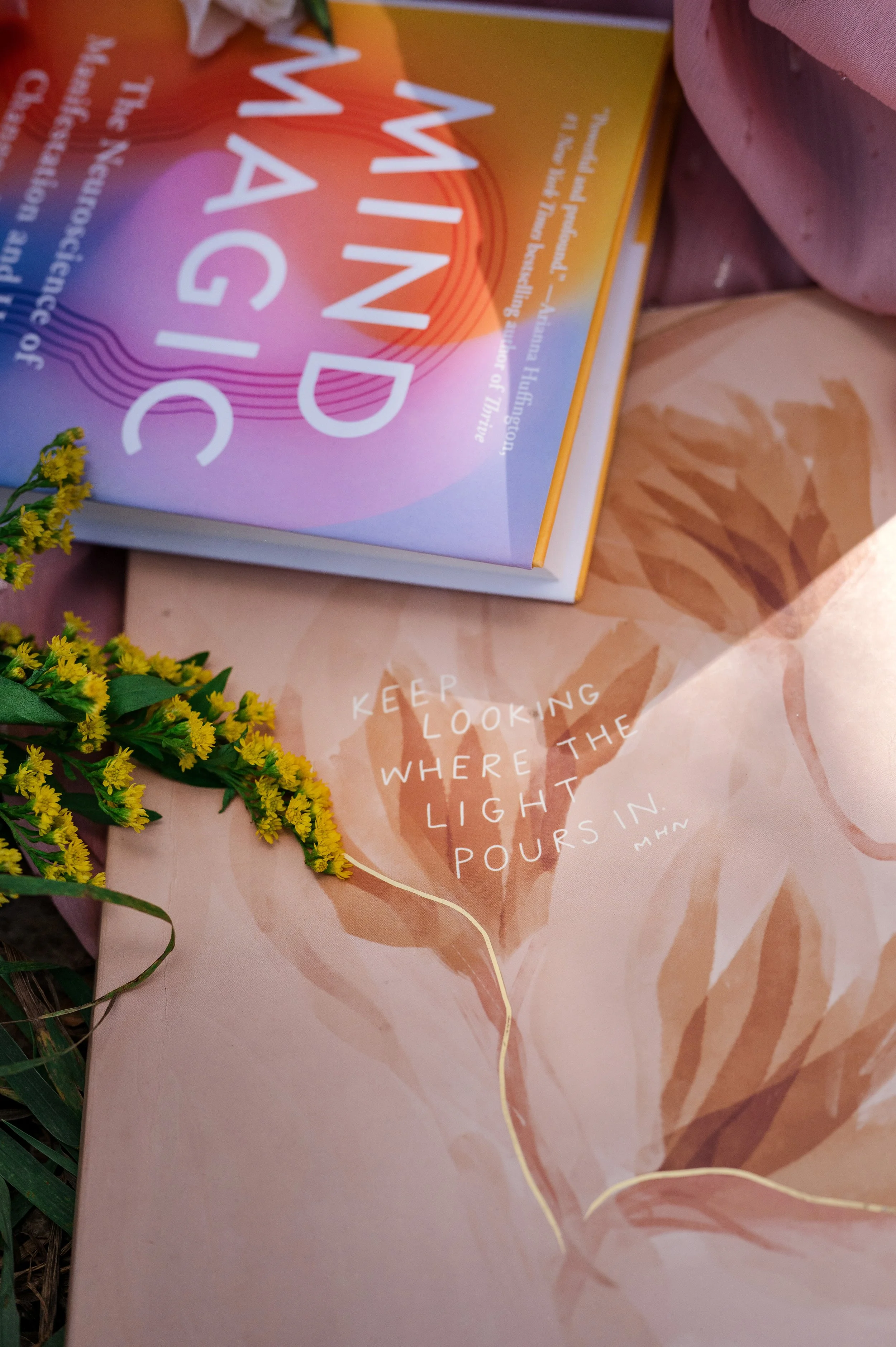 Close-up of a colorful book called "Mind Magic" on a floral-patterned journal with a motivational quote that says, "Keep looking where the light pours in." A sprig of yellow flowers is placed beside the book.