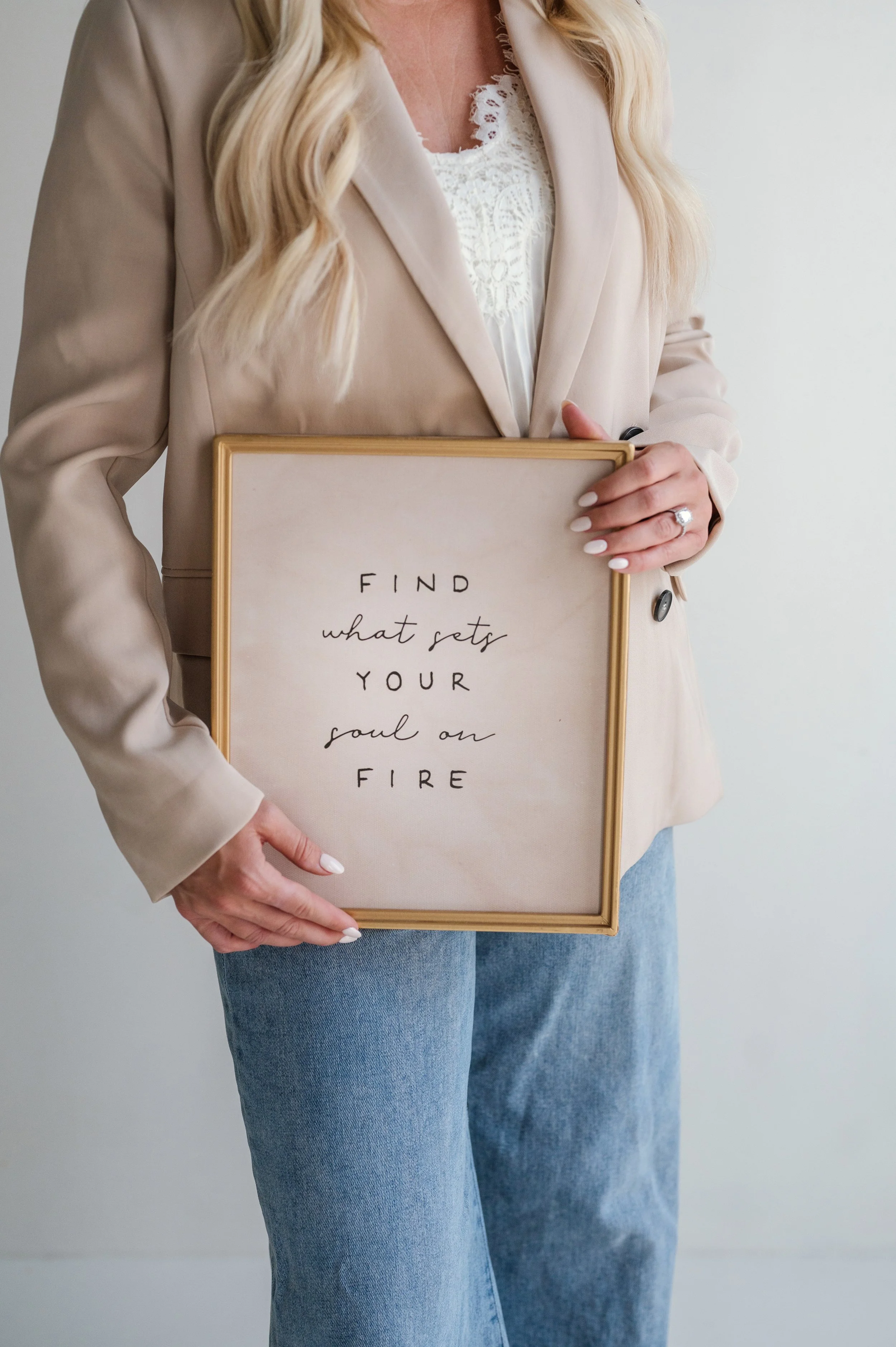 A woman with long blonde hair wearing a beige blazer and white lace top holding a framed sign with the text "FIND what sets YOUR soul on FIRE" written on it, standing against a plain background.