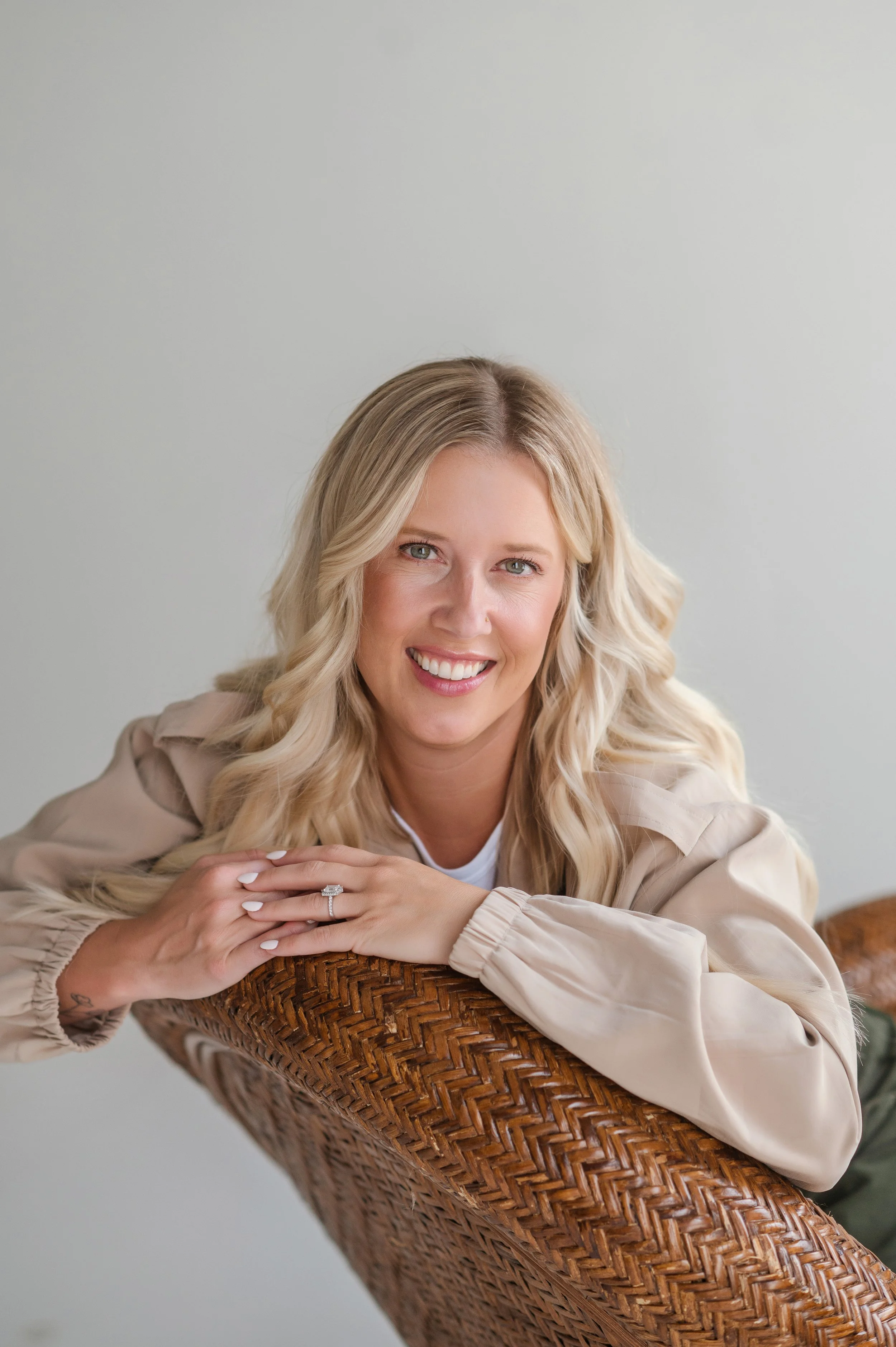 A woman with long blonde hair smiling, resting her arms on a woven basket, wearing a light beige jacket and a ring.